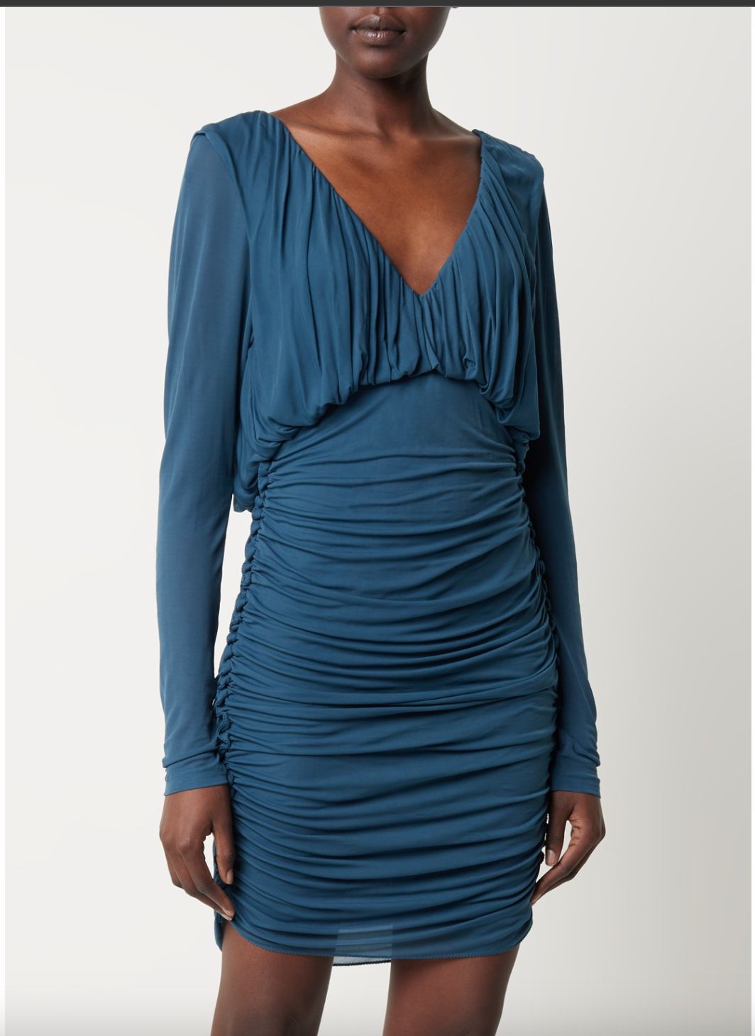 E-commerce shot of a model in a teal ruched long-sleeve mini dress with deep V-neckline, plain light backdrop