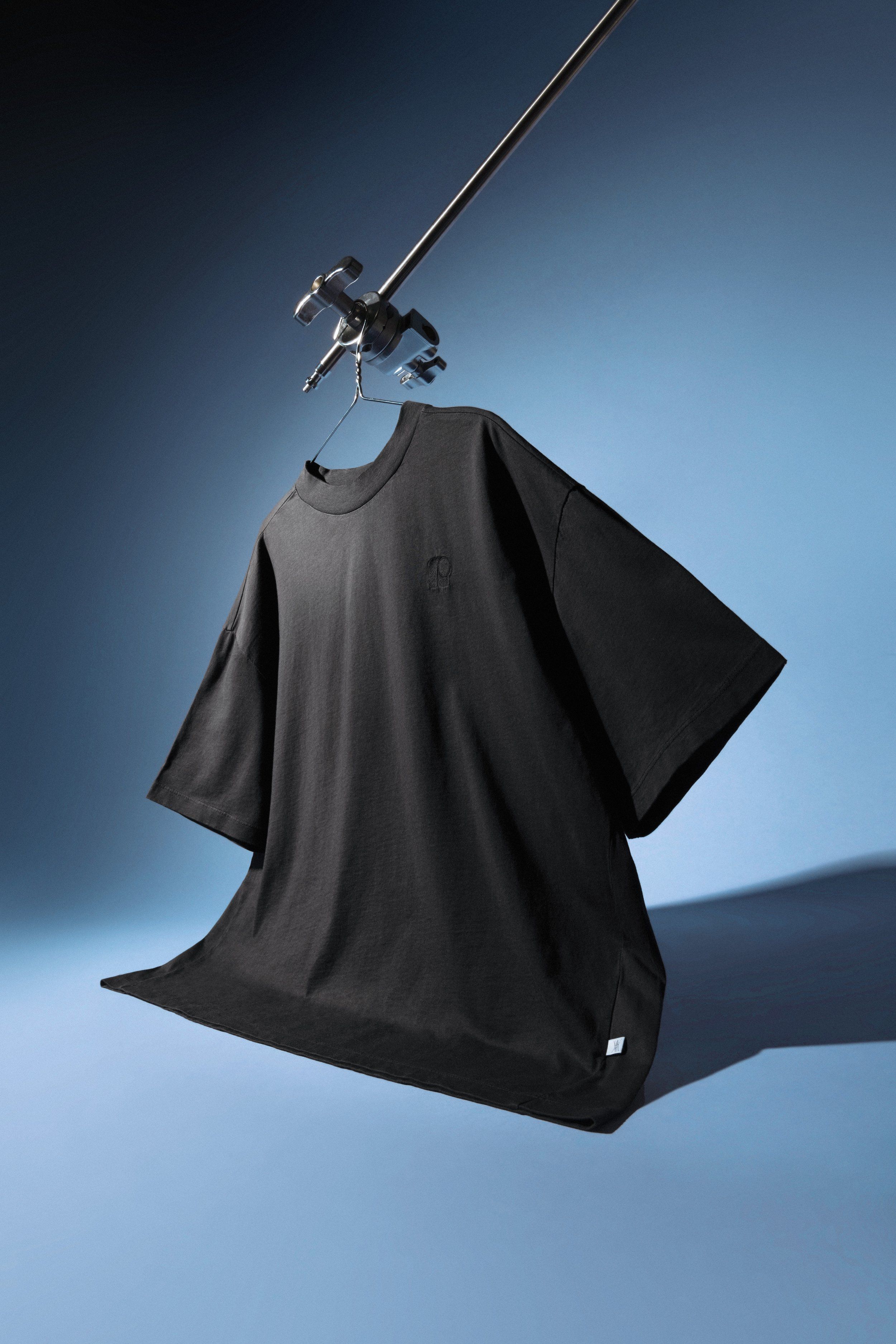 Oversized black t-shirt hanging from a studio boom arm against a blue gradient background, creative product shot