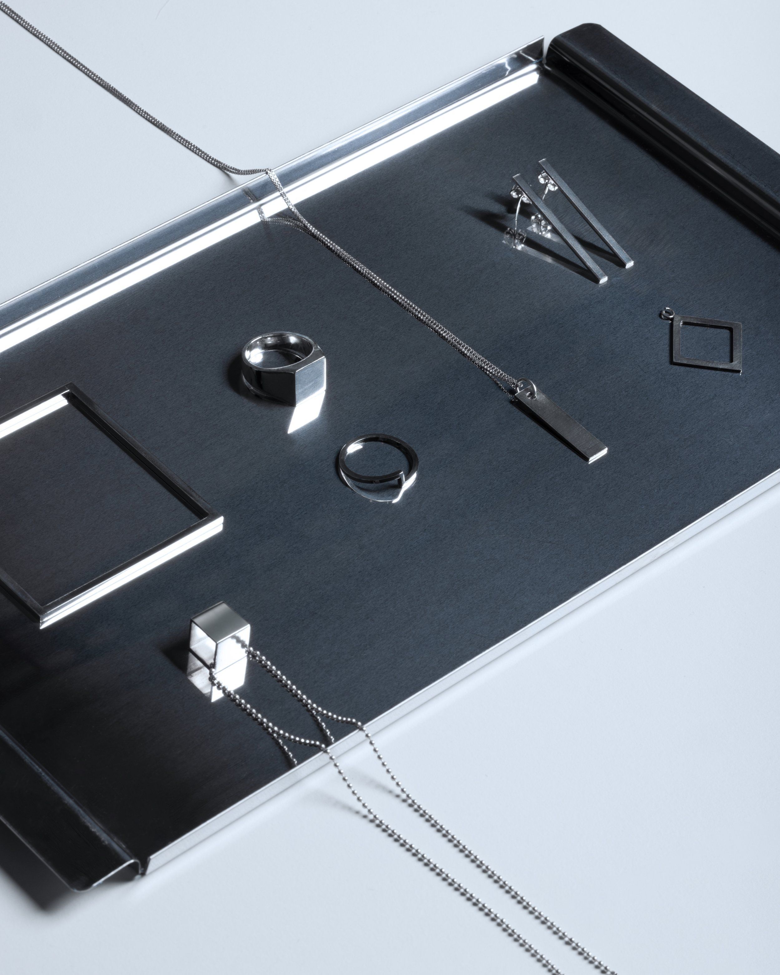 Silver rings, pendants, earrings and chains arranged on a dark brushed metal tray, flat-lay with cool reflected light