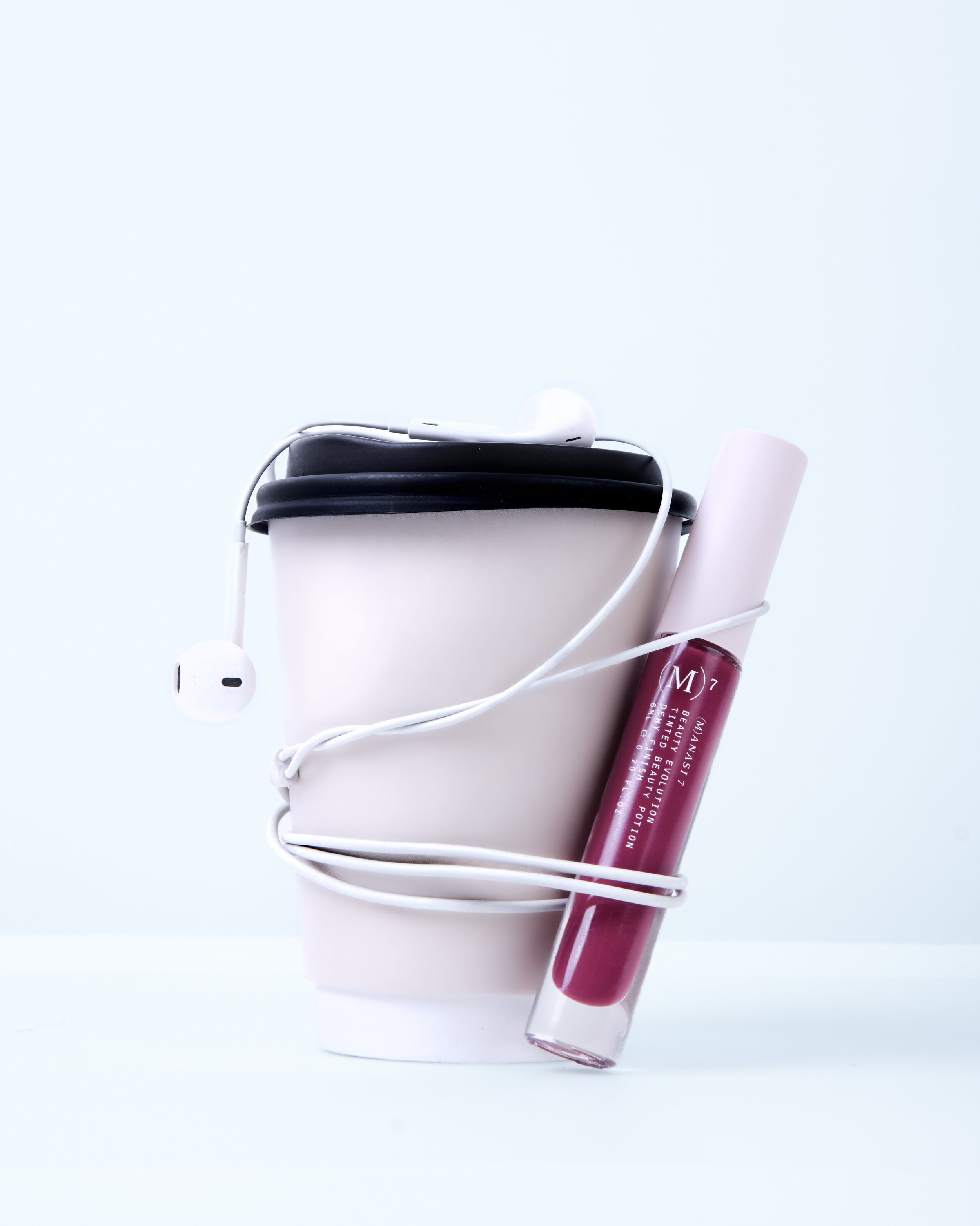 Burgundy lip gloss tube leaned on a takeaway coffee cup wrapped in white earphones, pale blue background
