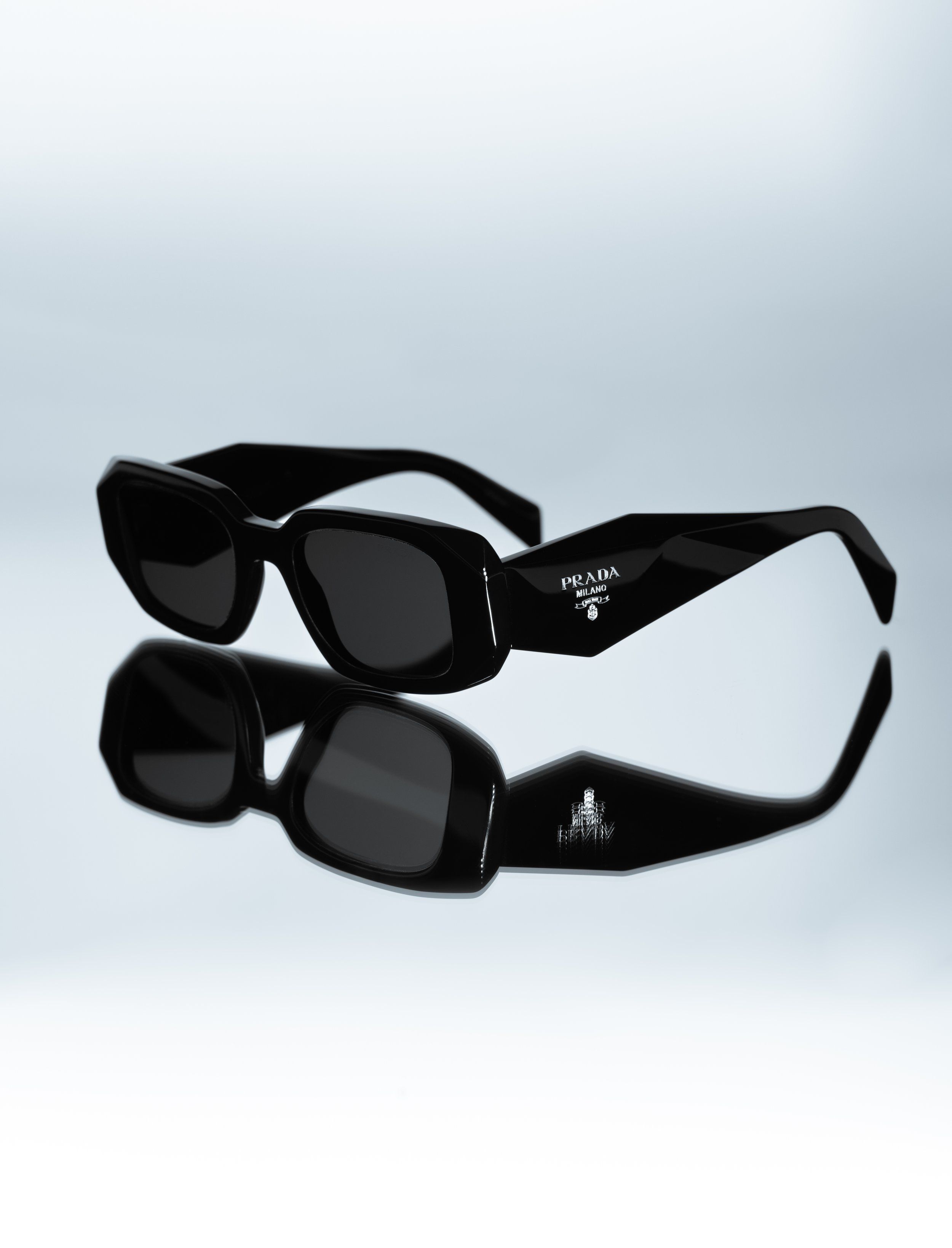 Black rectangular Prada sunglasses on a reflective surface, mirrored in the glossy floor against a soft grey backdrop