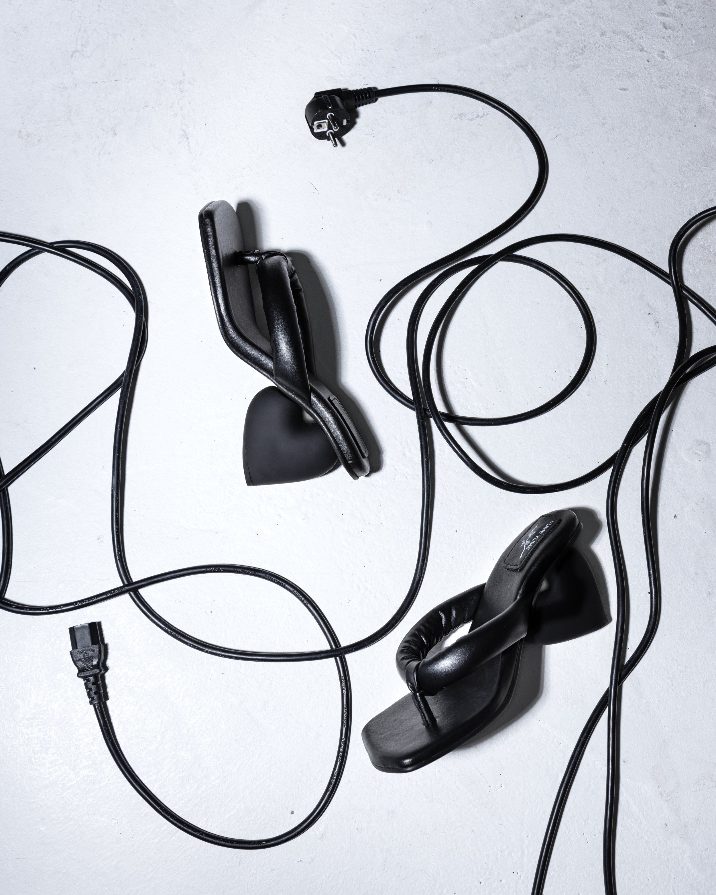 Black padded leather thong sandals scattered among black power cables on a pale concrete-look surface