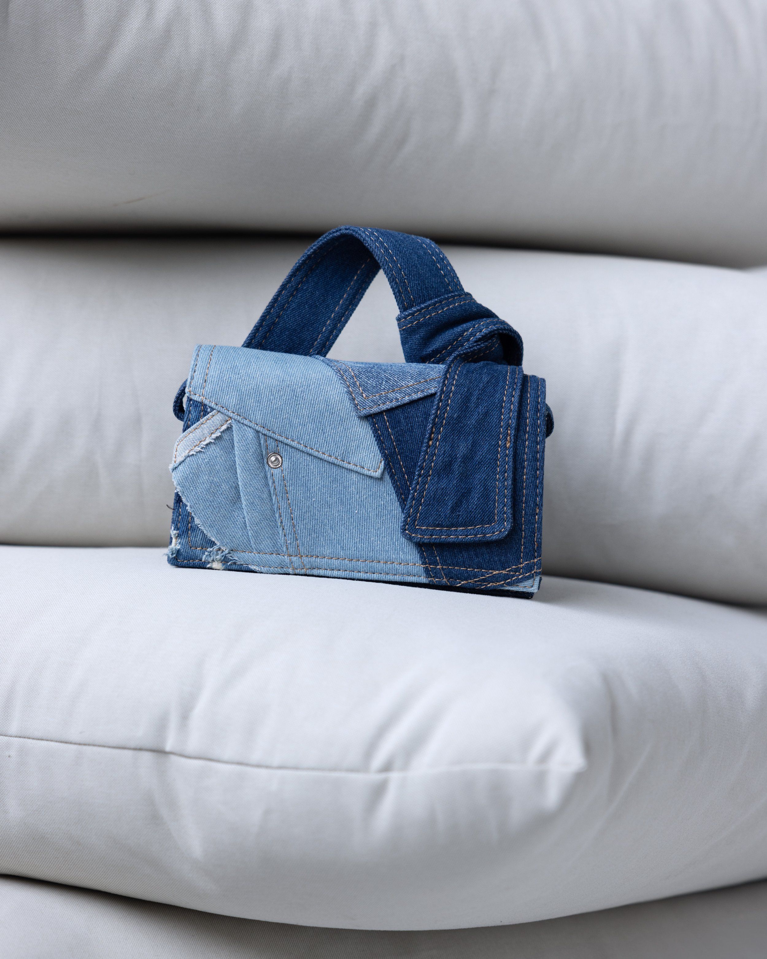 Small patchwork denim handbag with a knotted handle resting on a stack of white pillows