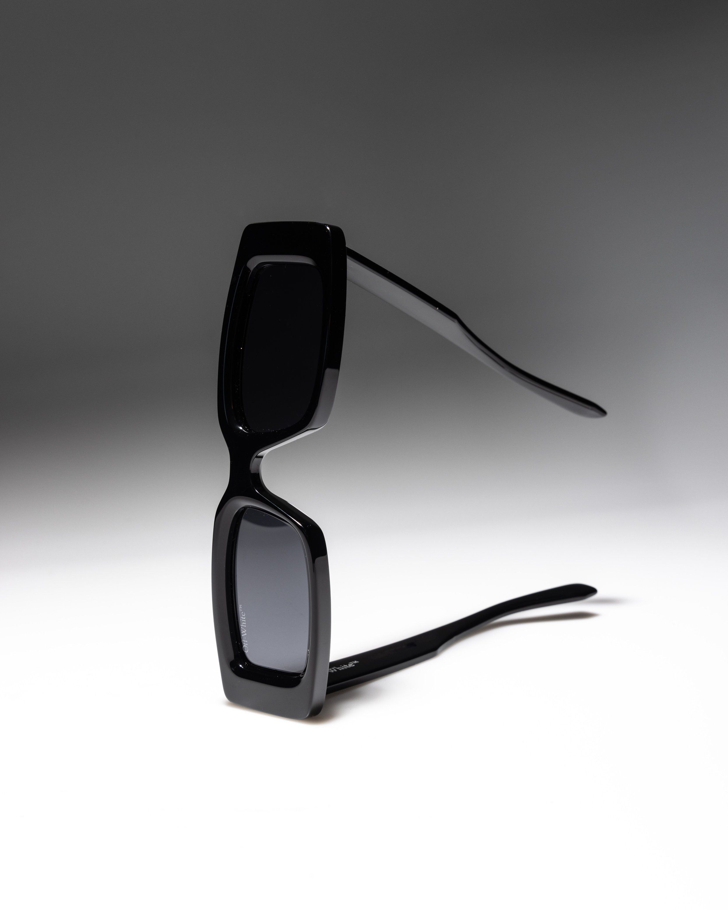 Black chunky rectangular sunglasses balanced upright on a white surface with a dark grey gradient background