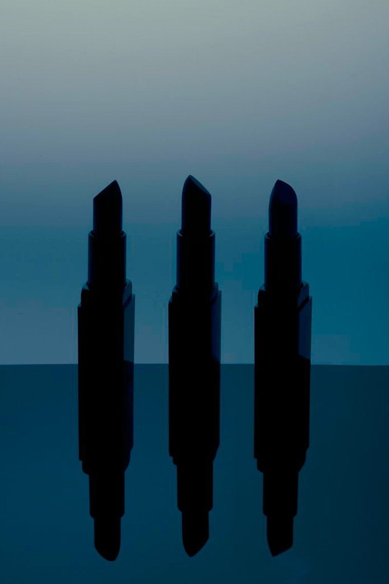 Three black lipsticks in silhouette on a reflective surface against a blue-teal gradient background