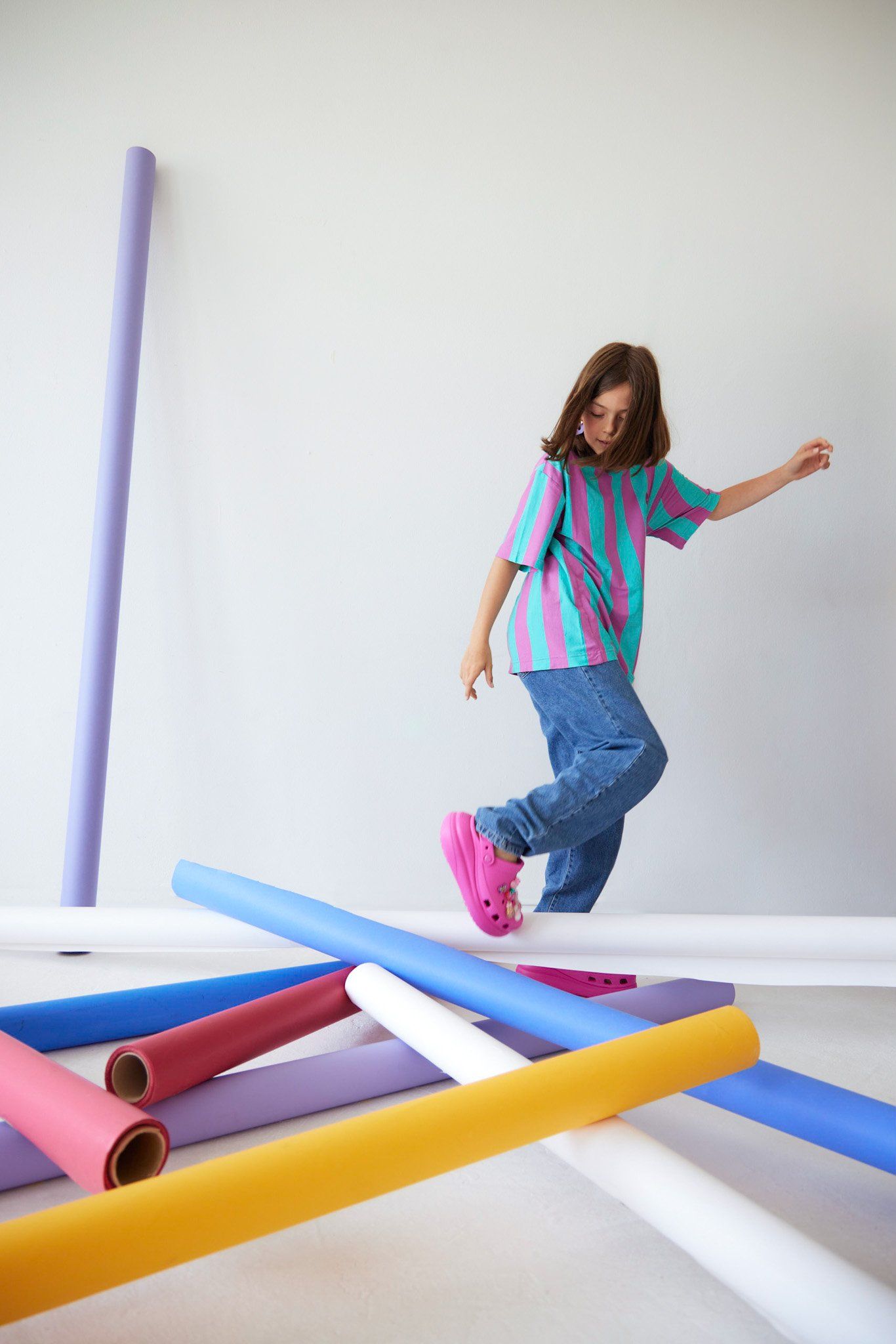 Child model in a striped turquoise and pink tee, jeans and pink clogs walking among colourful paper rolls on white