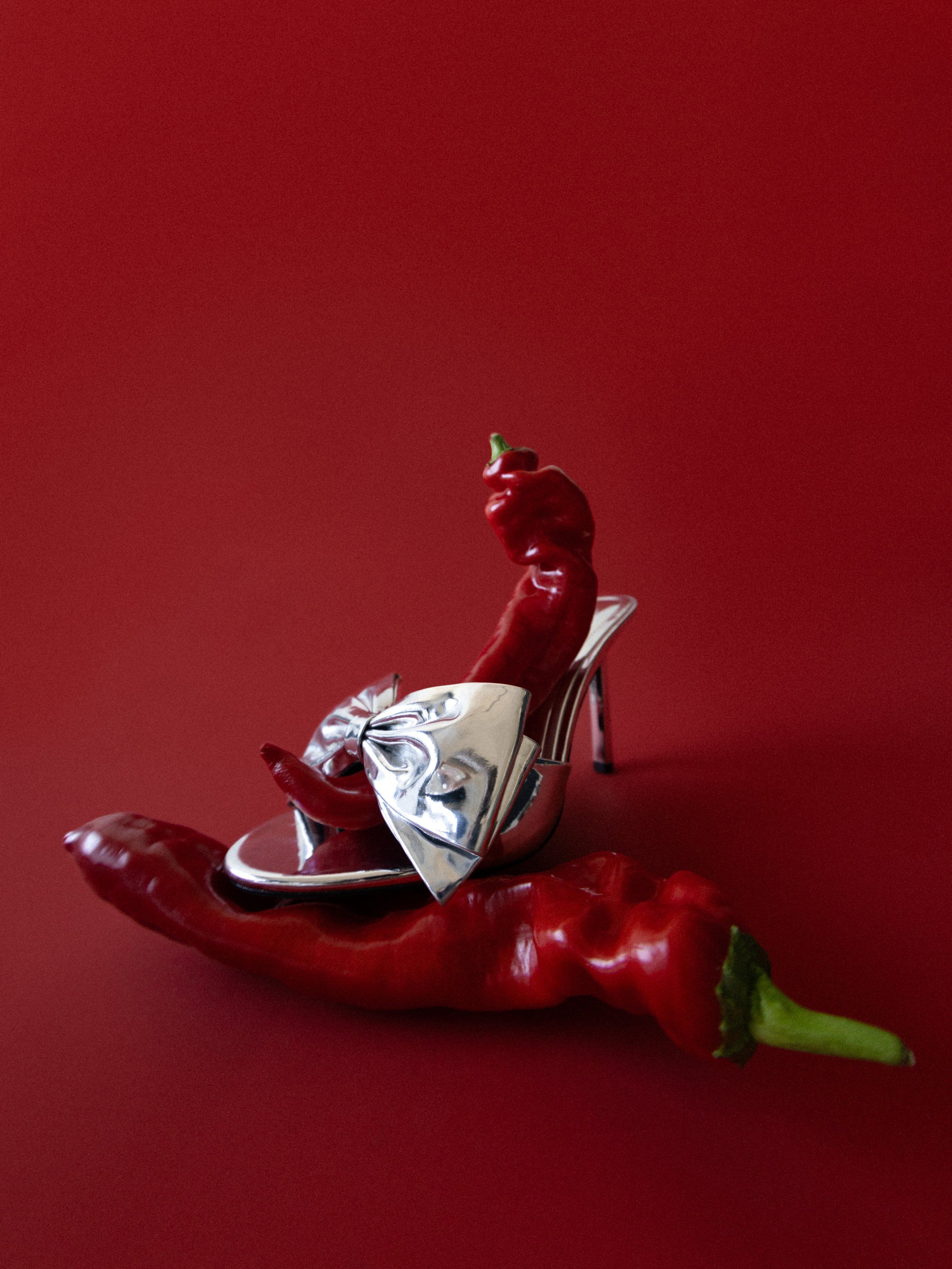 Silver bow-front high-heeled mule among red chili peppers on a deep red background
