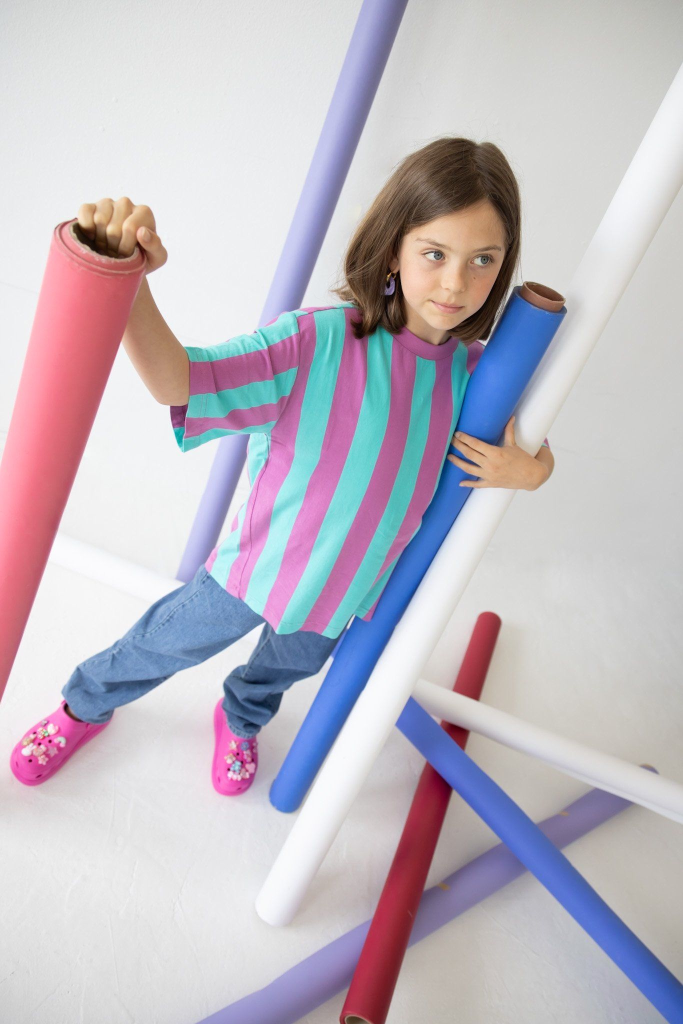 Child model in a turquoise and pink striped tee and pink clogs posed among pink, blue and white paper rolls