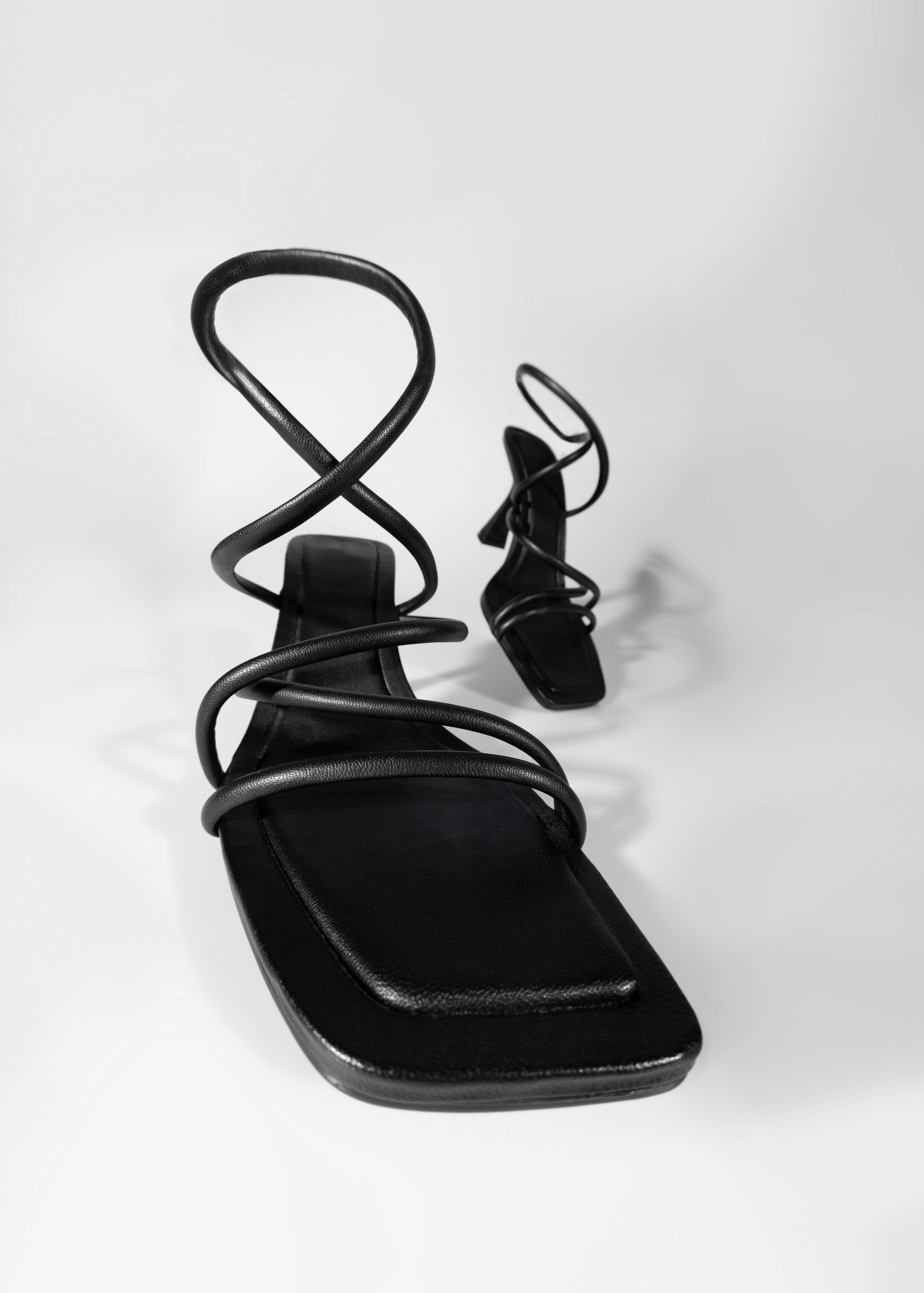 Black strappy square-toe sandals with curling ankle straps on a pale grey background, shown from two angles