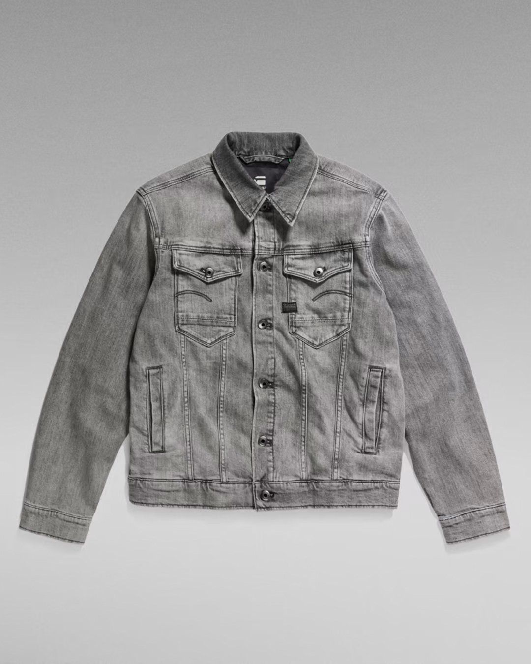 Grey washed denim trucker jacket with chest pockets, flat lay on grey gradient background, G-Star RAW