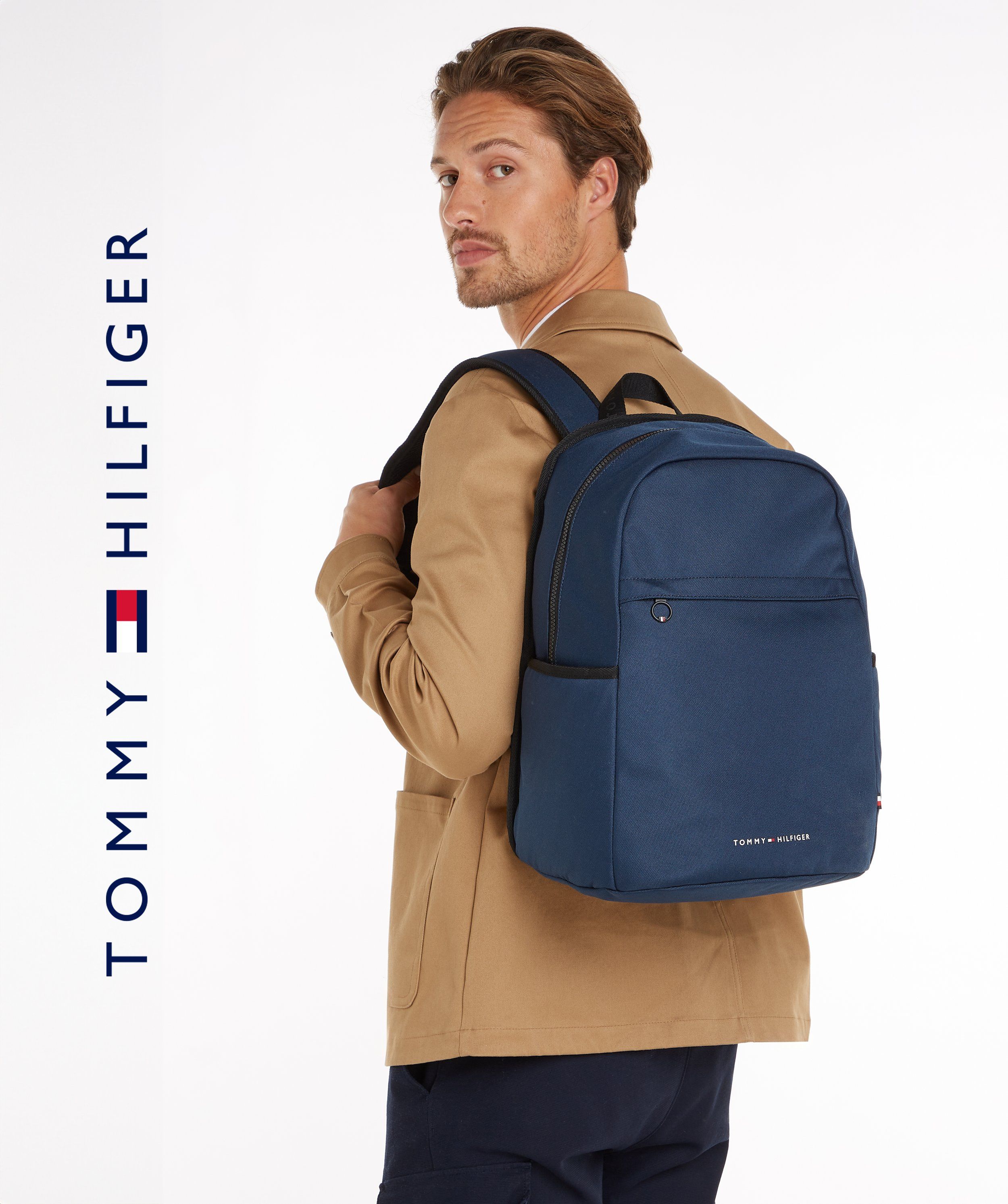 Tommy Hilfiger logo beside a male model in a camel jacket carrying a navy backpack, glancing back, white background