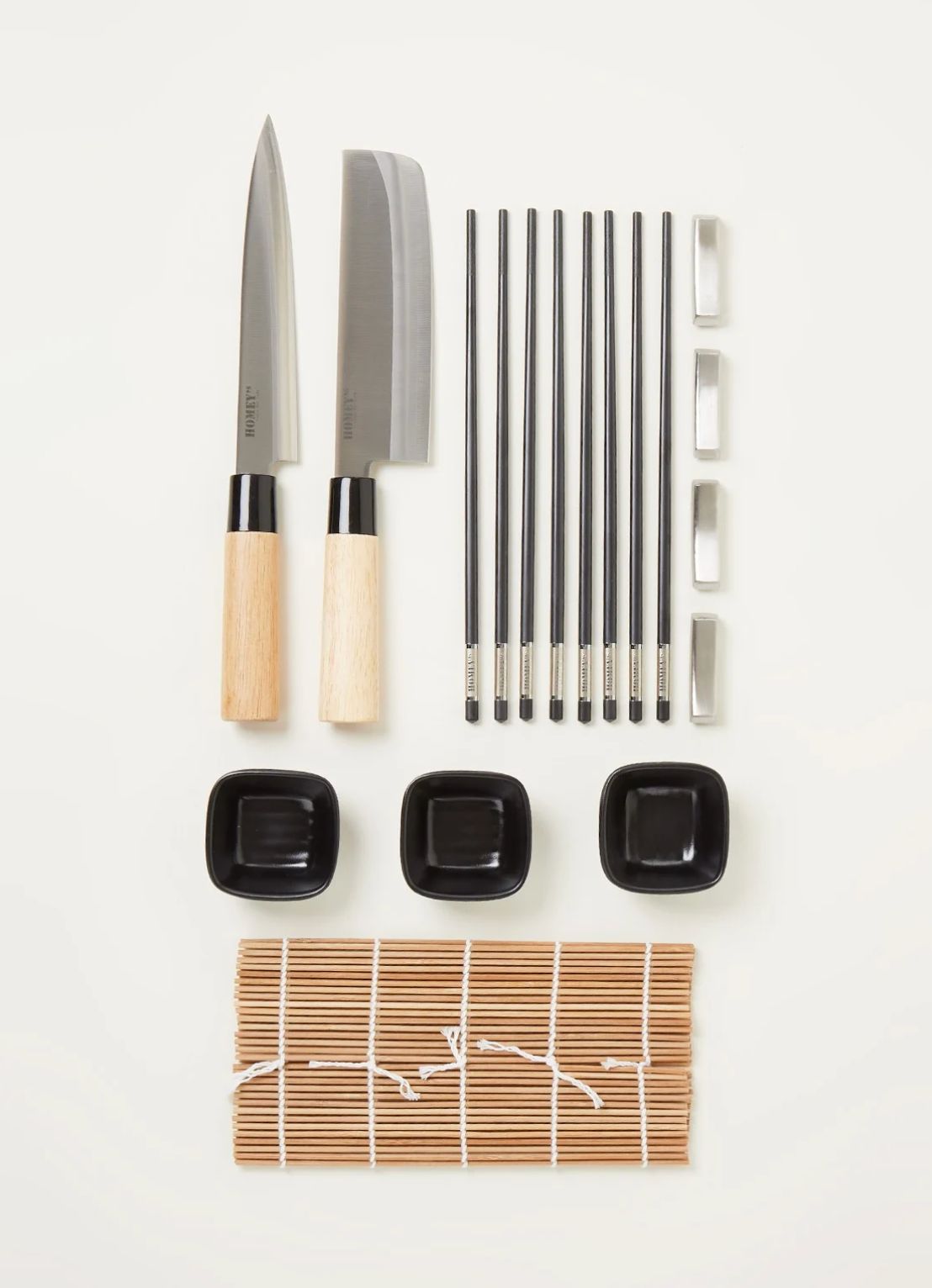 Flat lay of two Japanese knives, chopsticks, rests, black dishes and a bamboo rolling mat on off-white