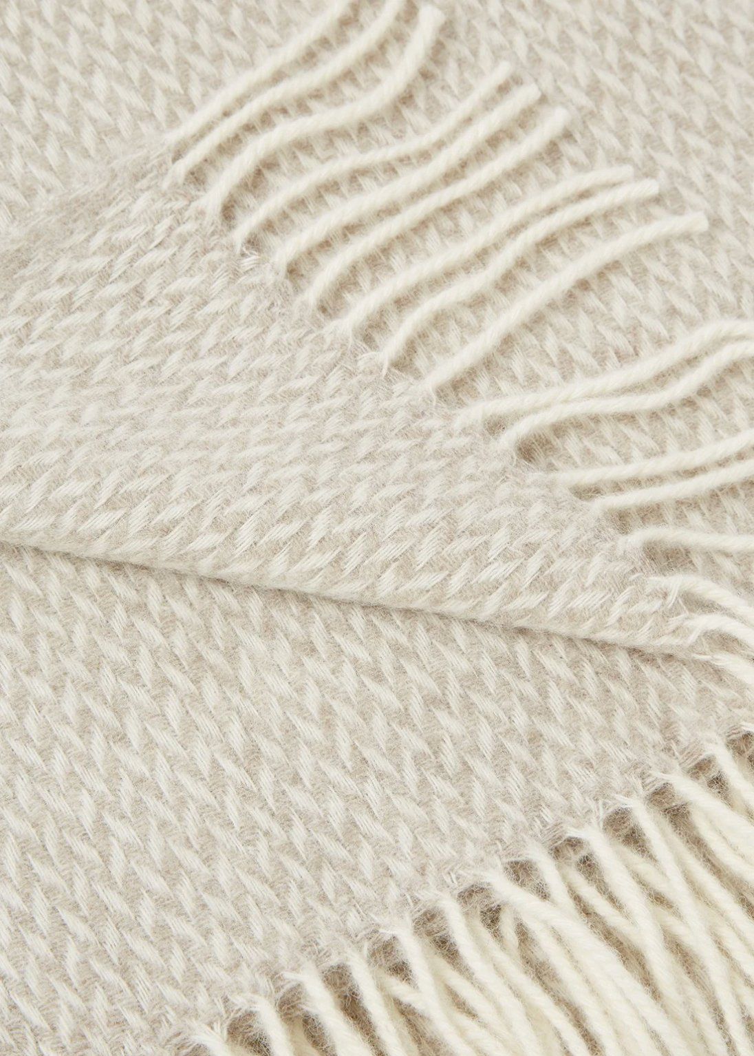 Close-up of beige herringbone wool throw with cream fringe, texture detail on light background