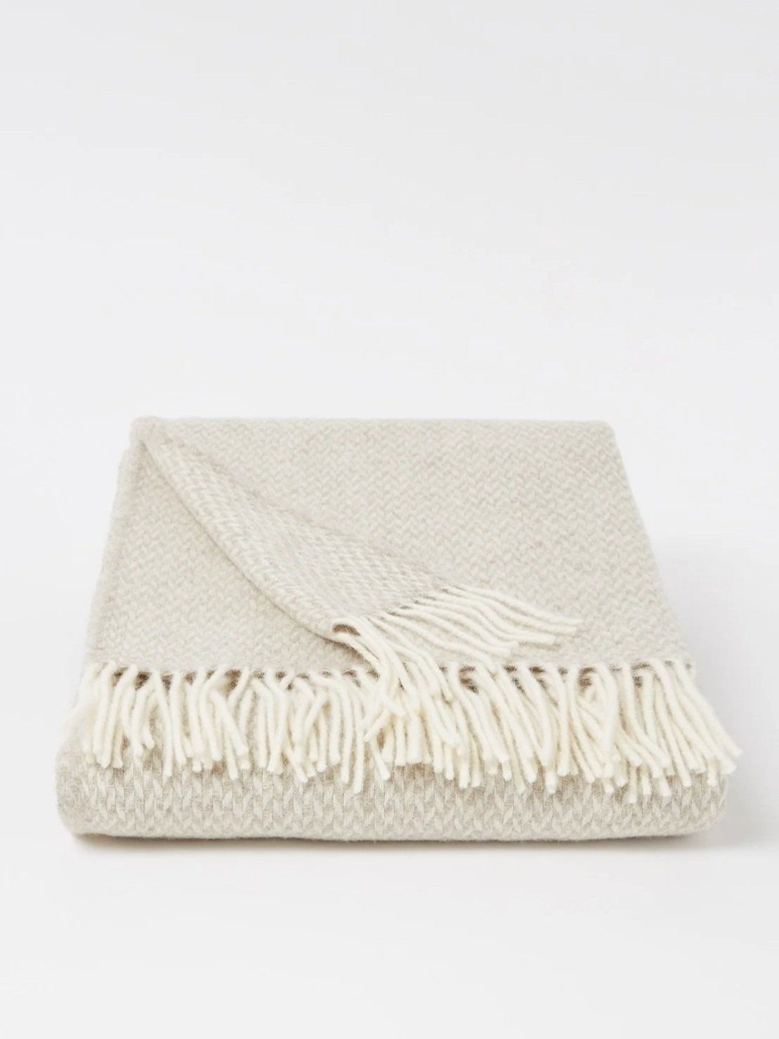 Folded beige wool throw with fringed edge on a light grey studio background