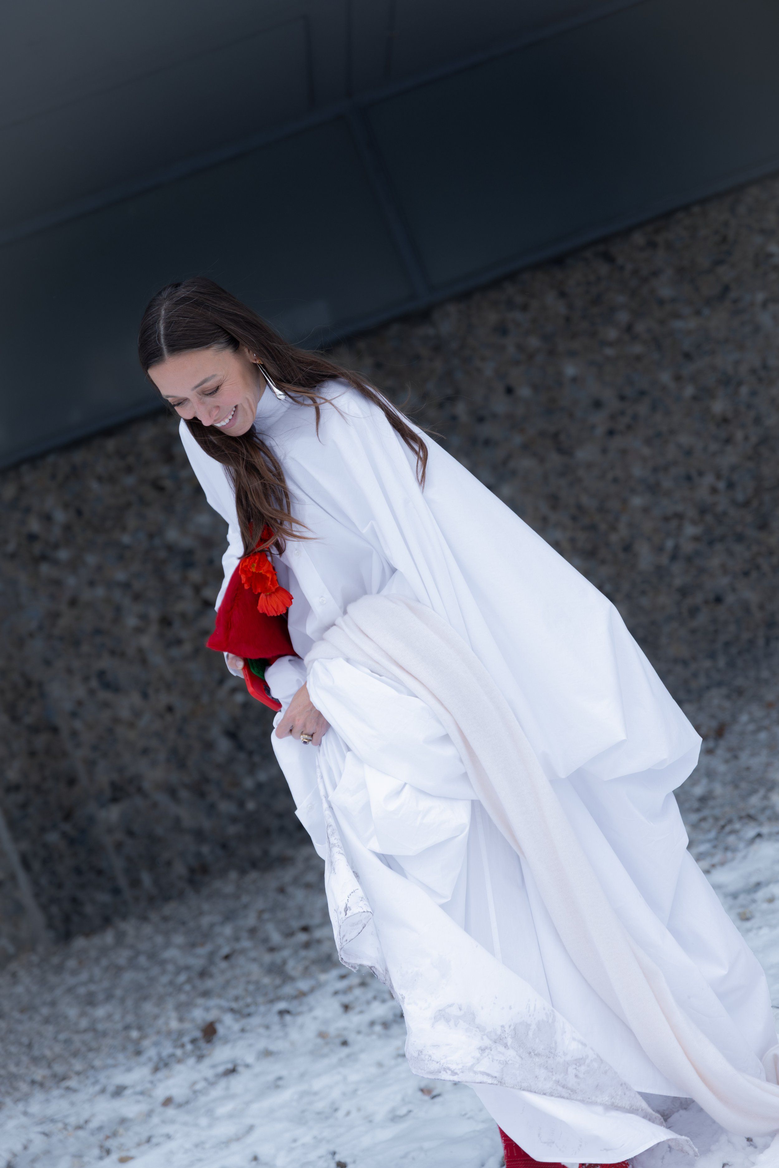Woman in a flowing white cape-like dress lifting the hem, red bag at her side, on a snowy pavement