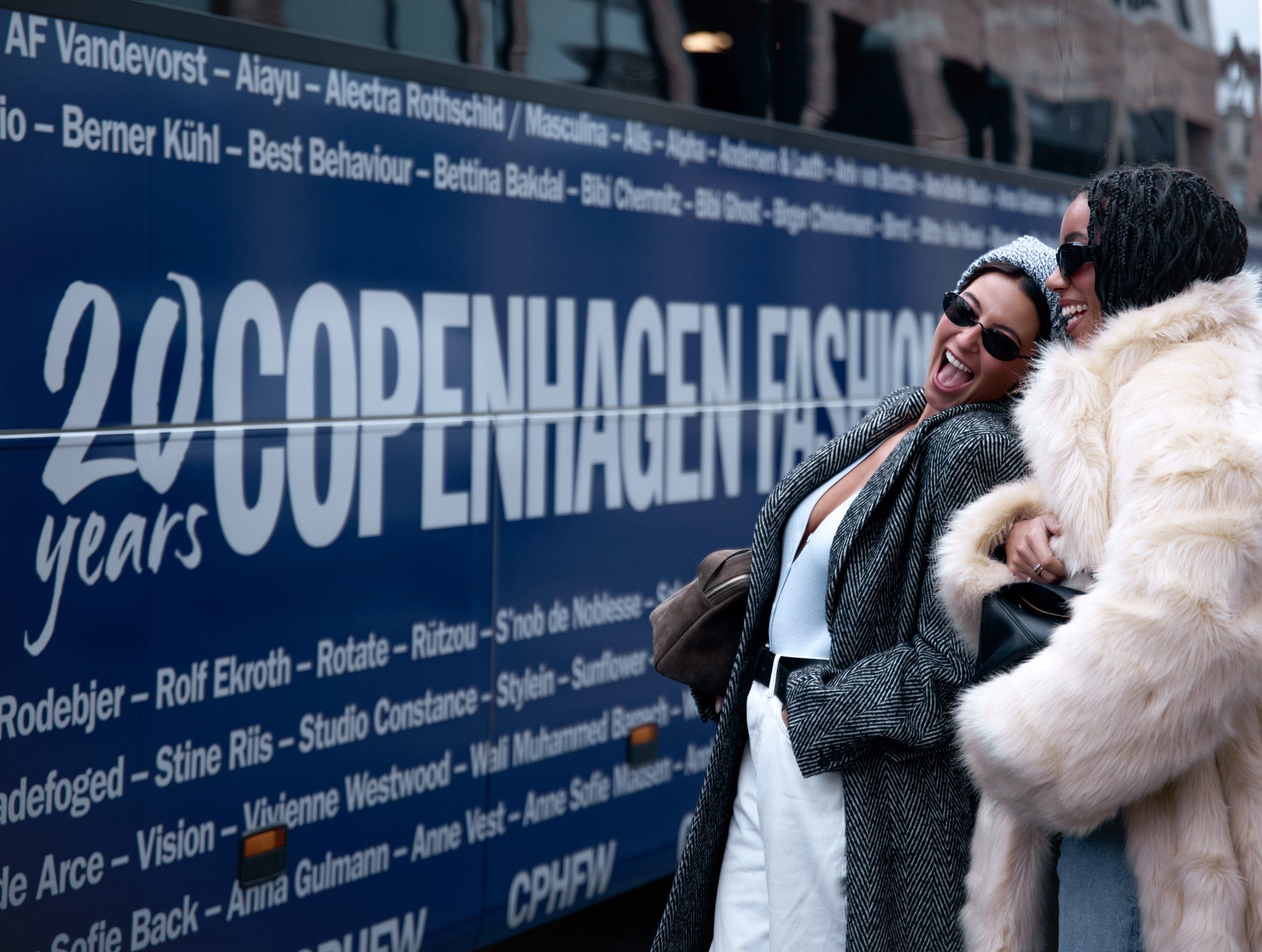 Copenhagen Fashion Week street style: two guests laughing beside a blue CPHFW bus, one in a cream fur coat