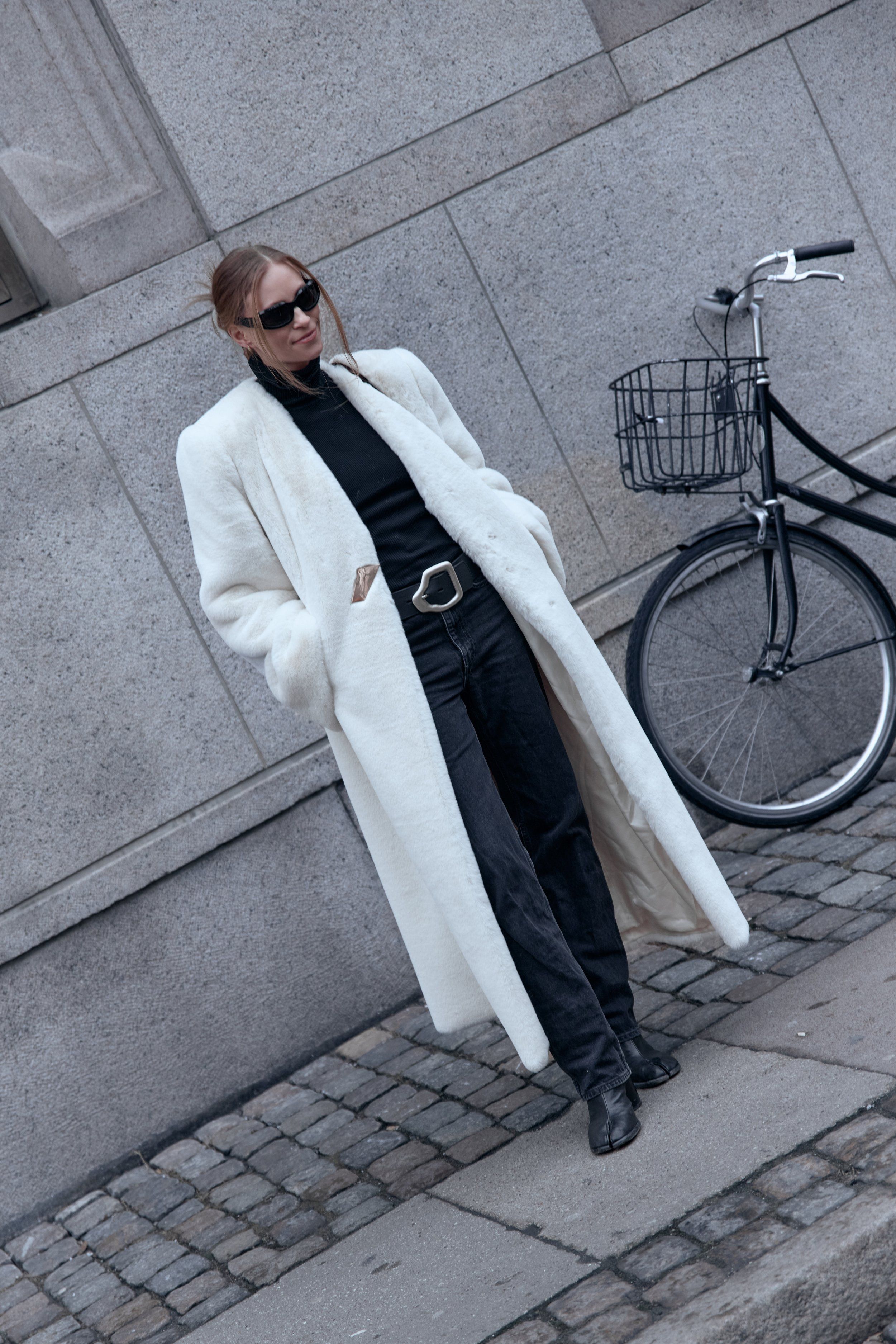 Woman in a long white coat, black top, dark jeans and sunglasses leaning on a stone wall beside a bicycle