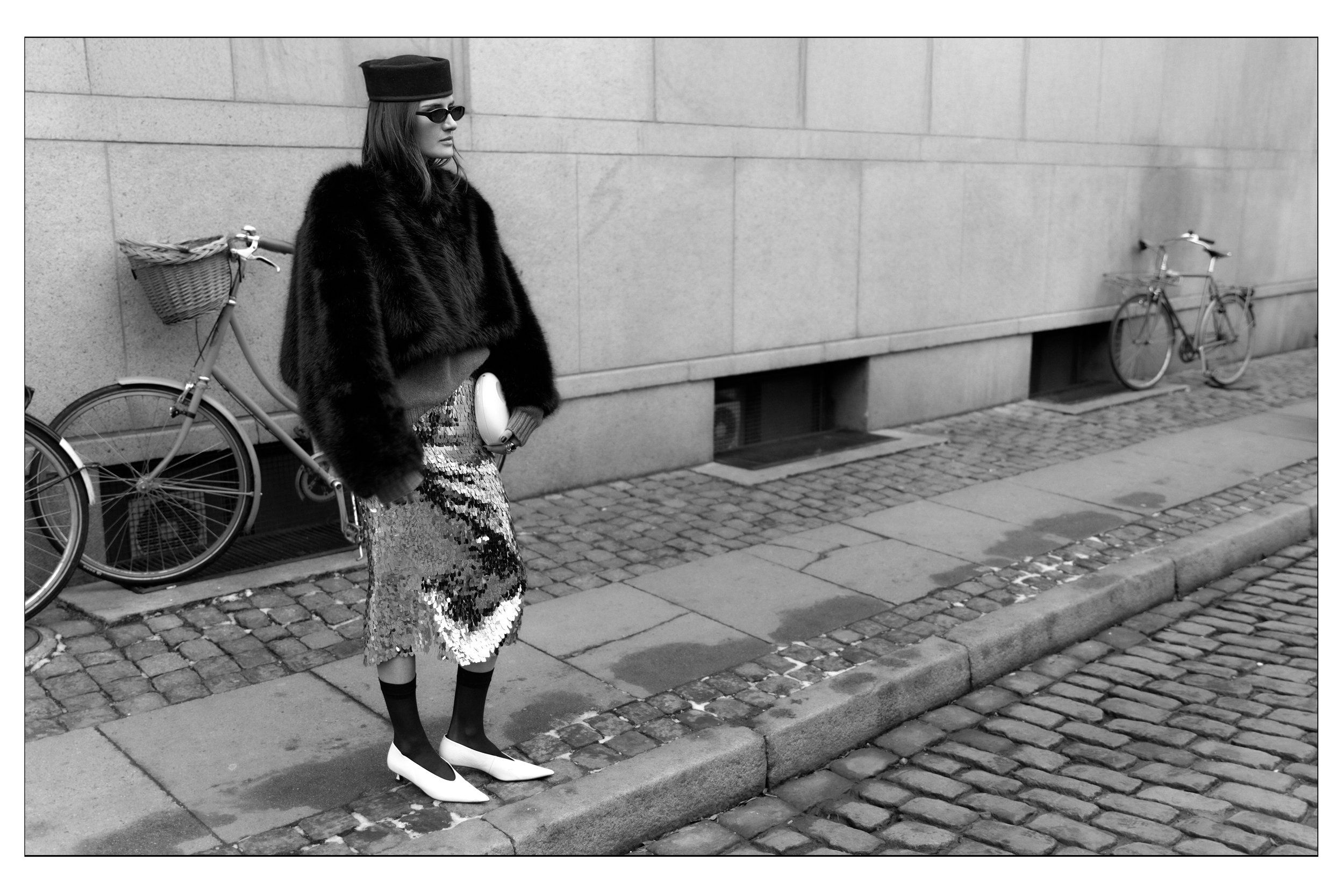 Black-and-white shot of a woman in a black fur jacket, bucket hat and patterned skirt on a cobbled street
