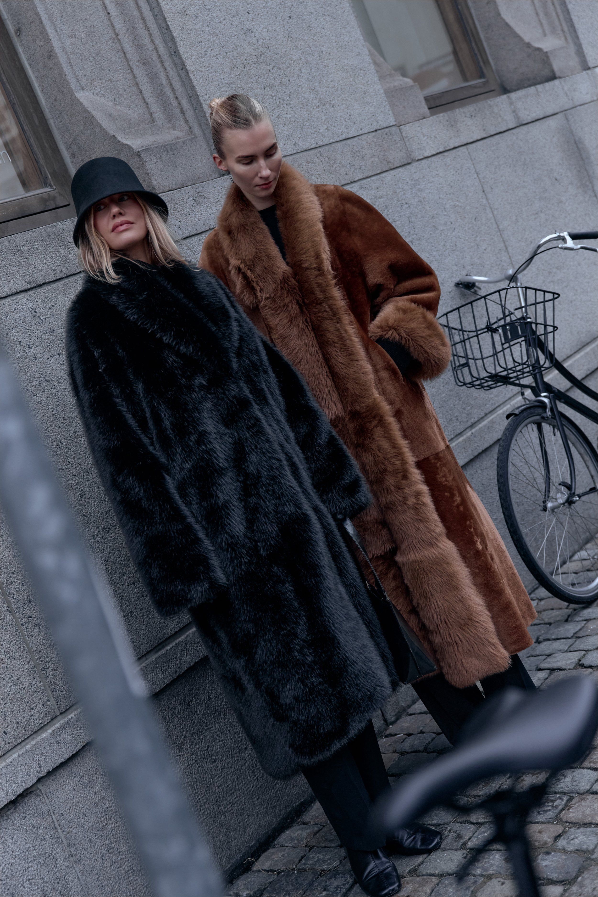 Two women in long coats, one black faux fur and one brown shearling, beside a bicycle with a basket