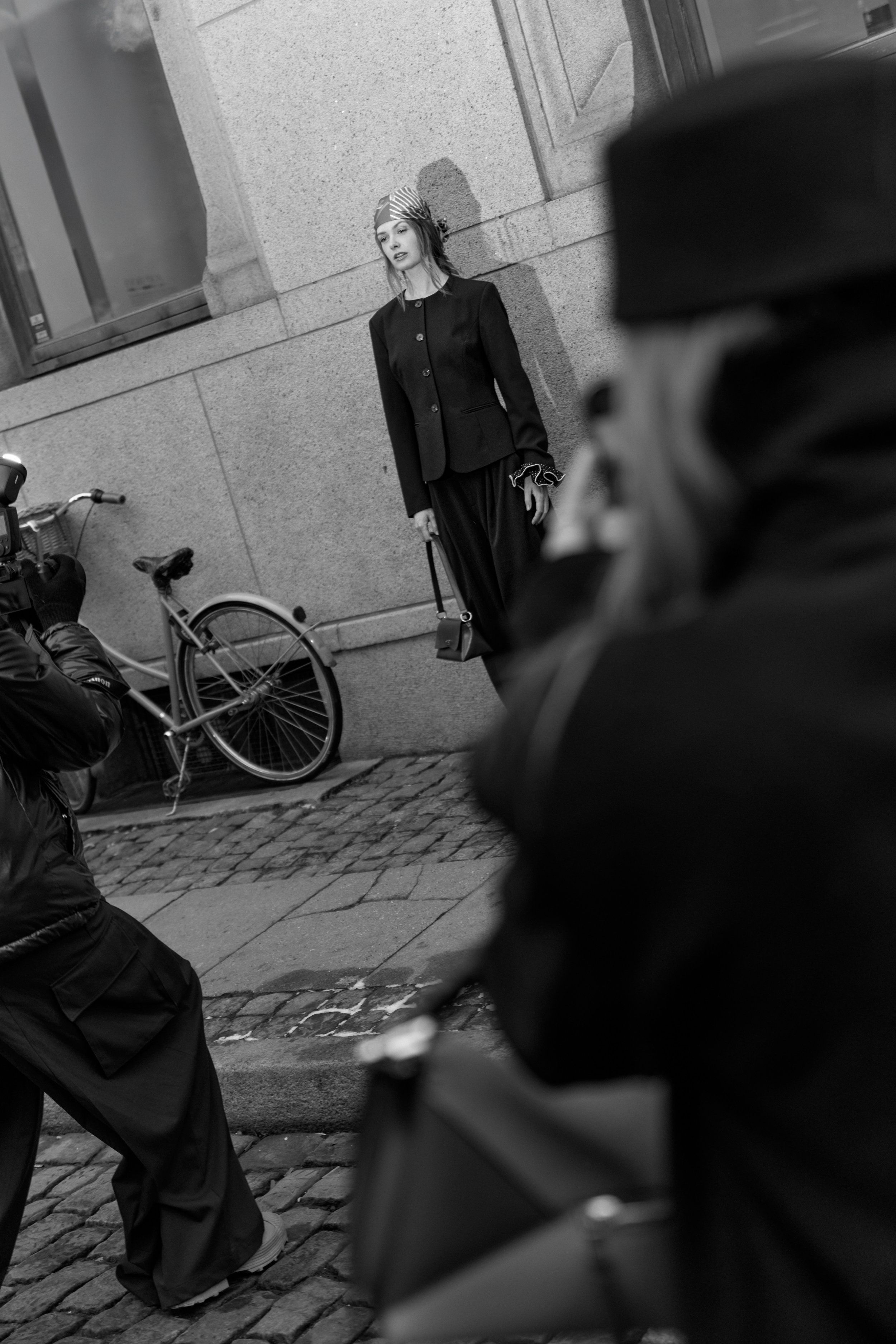 Black-and-white street scene with a woman in a dark jacket and skirt by a bicycle, a photographer's hat in foreground