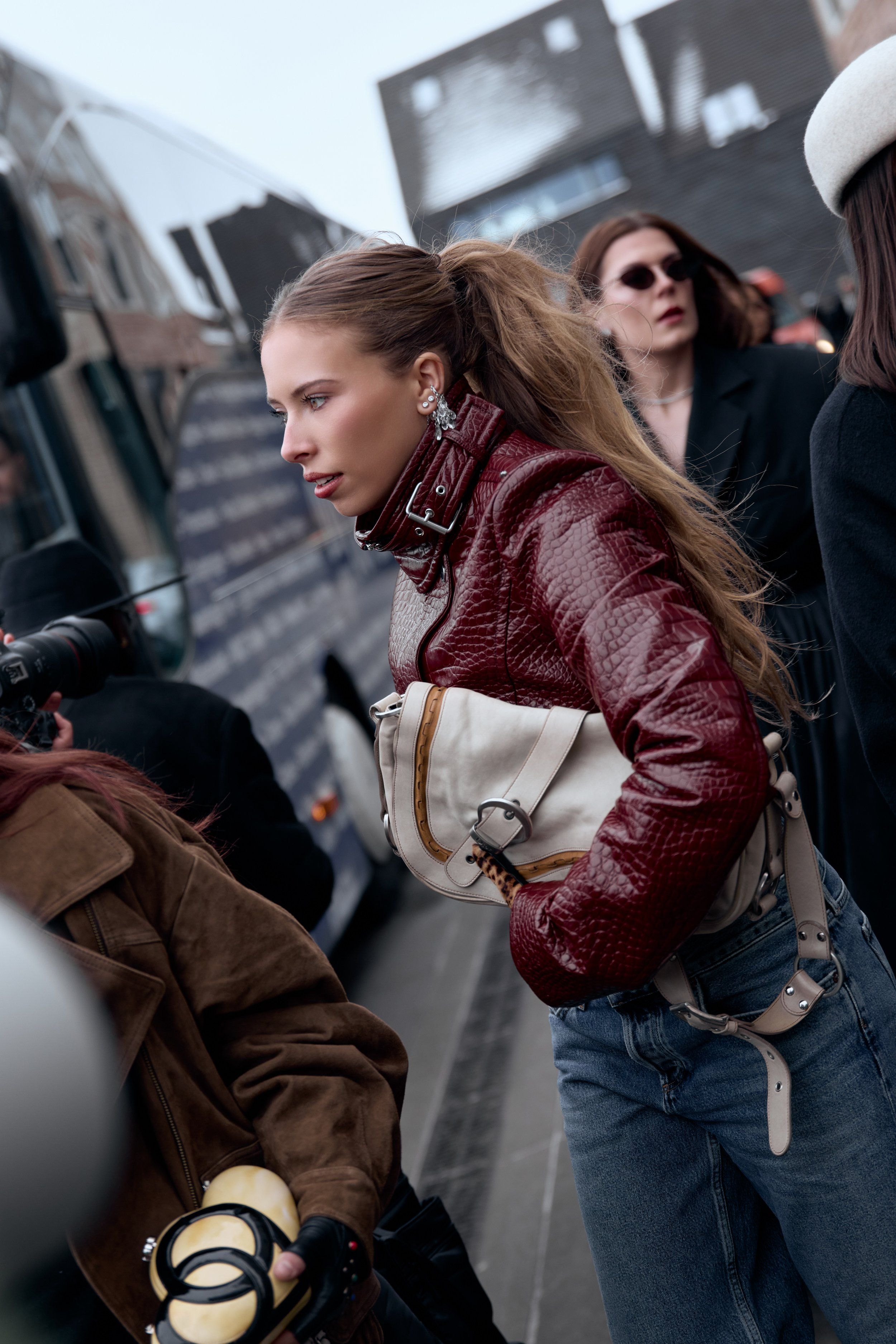 Woman in a burgundy croc-effect jacket with a cream belt bag and jeans, walking past the festival bus