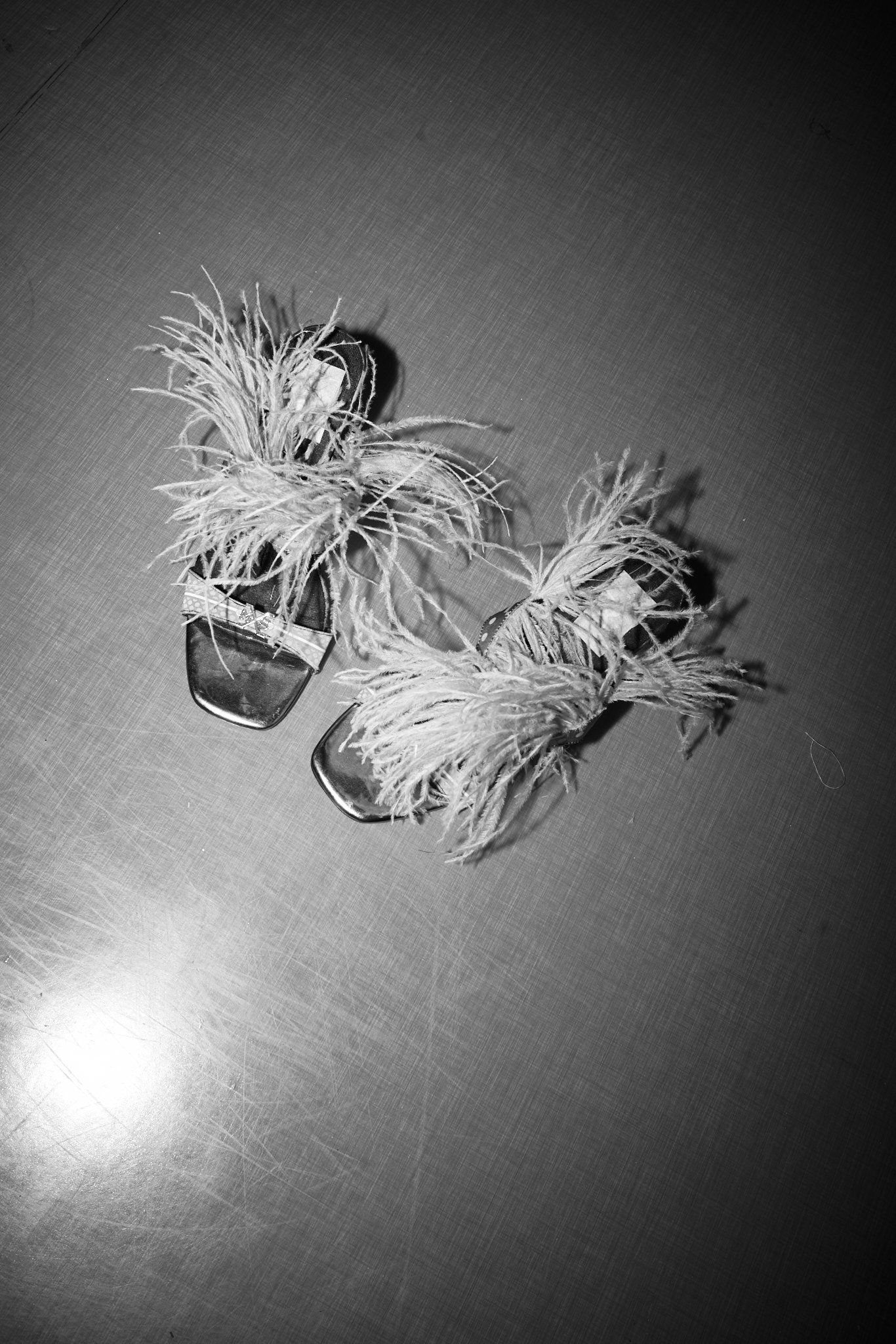 Black-and-white overhead shot of flat sandals trimmed with feathers on a reflective surface