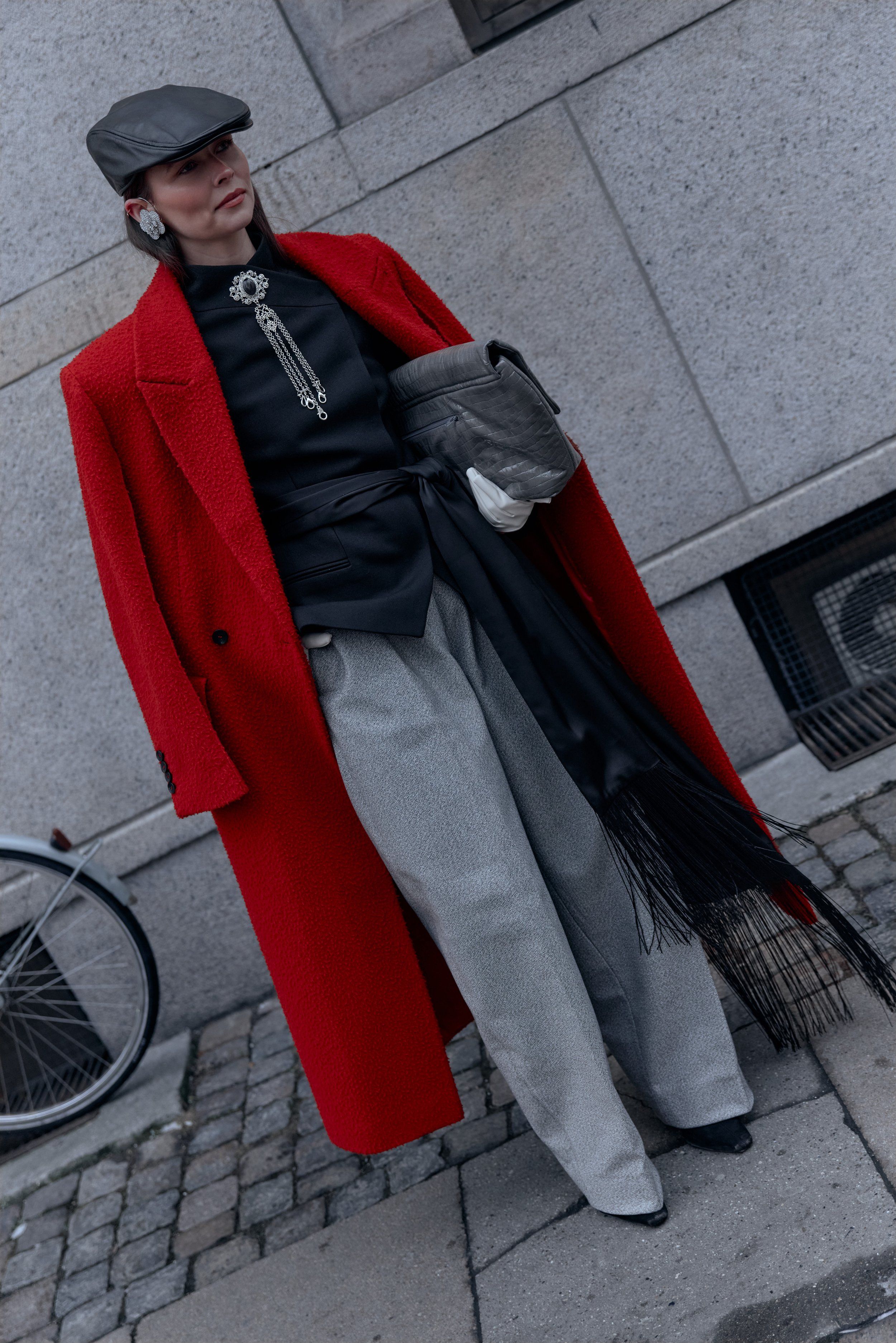 Woman in a long red coat, flat cap, black top with brooch and grey wide trousers, holding a fringed scarf