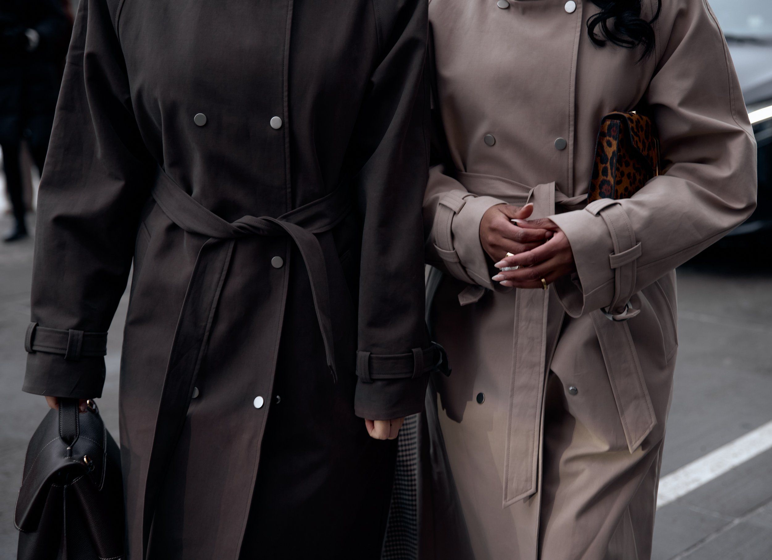 Two guests in belted trench coats, one dark brown and one beige, walking close together
