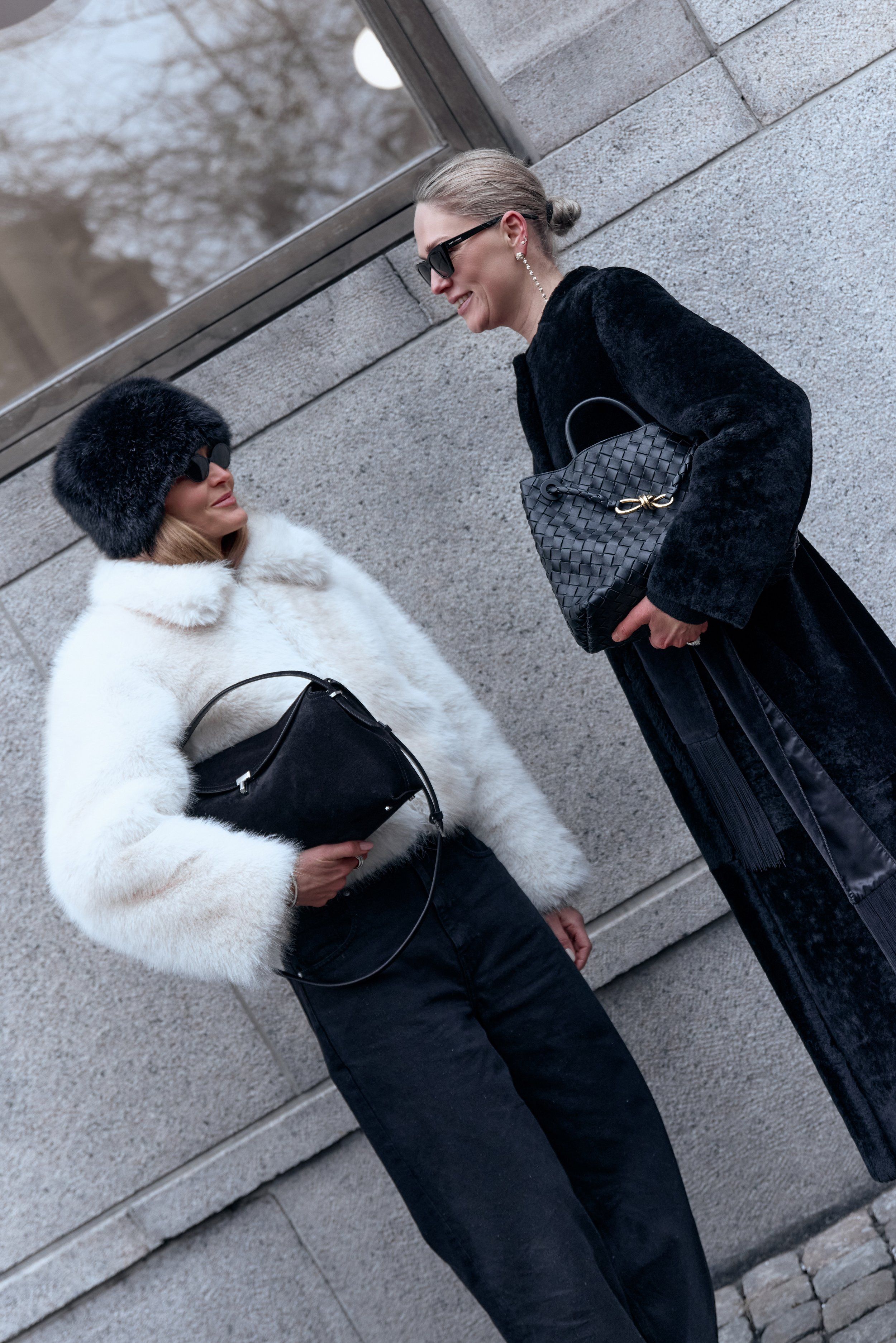 Two guests talking on a snowy street, one in a white fur jacket and black fur hat, one in a long black coat