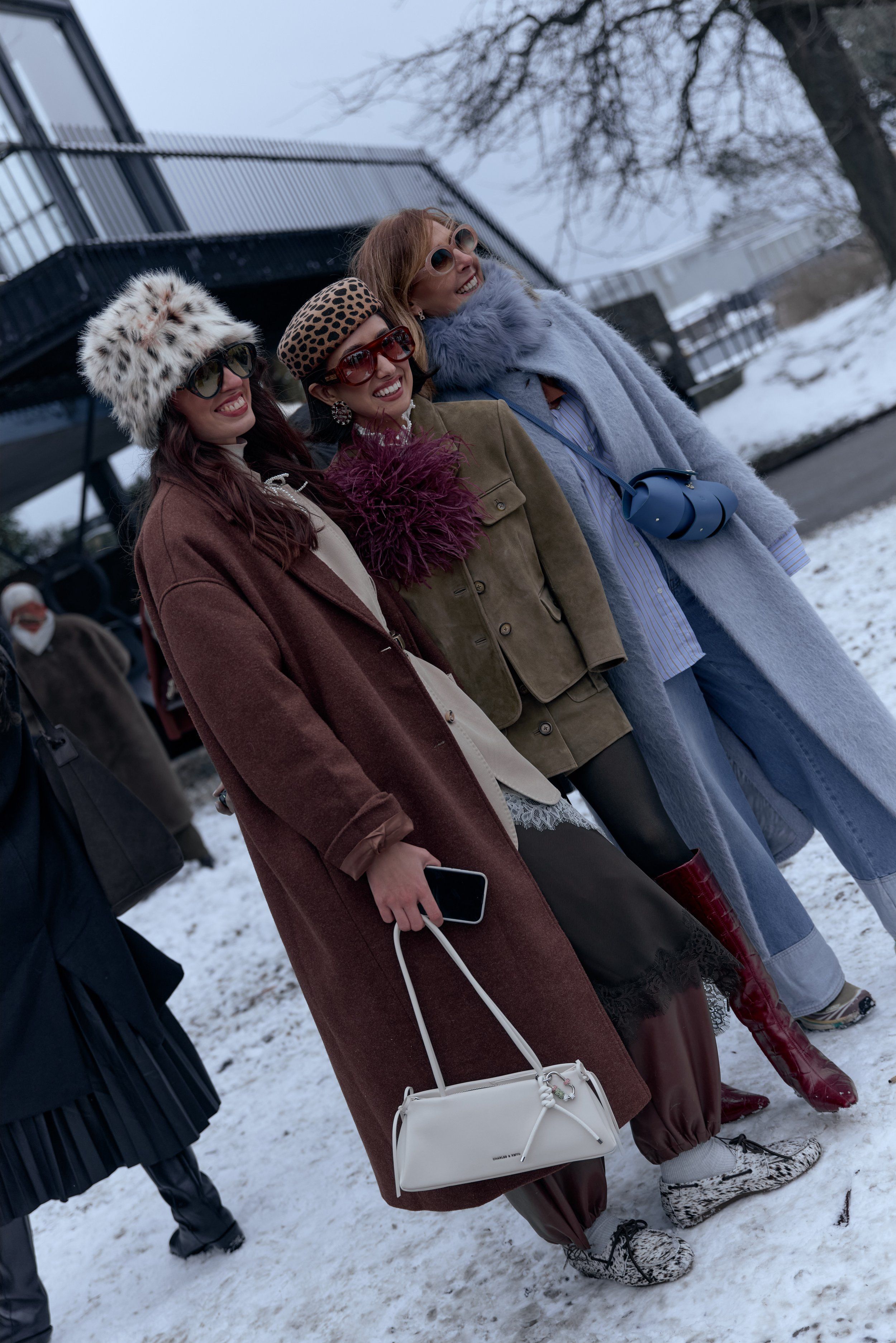 Three guests posing in the snow in patterned bucket hats, brown and blue coats, a feather collar and a white bag