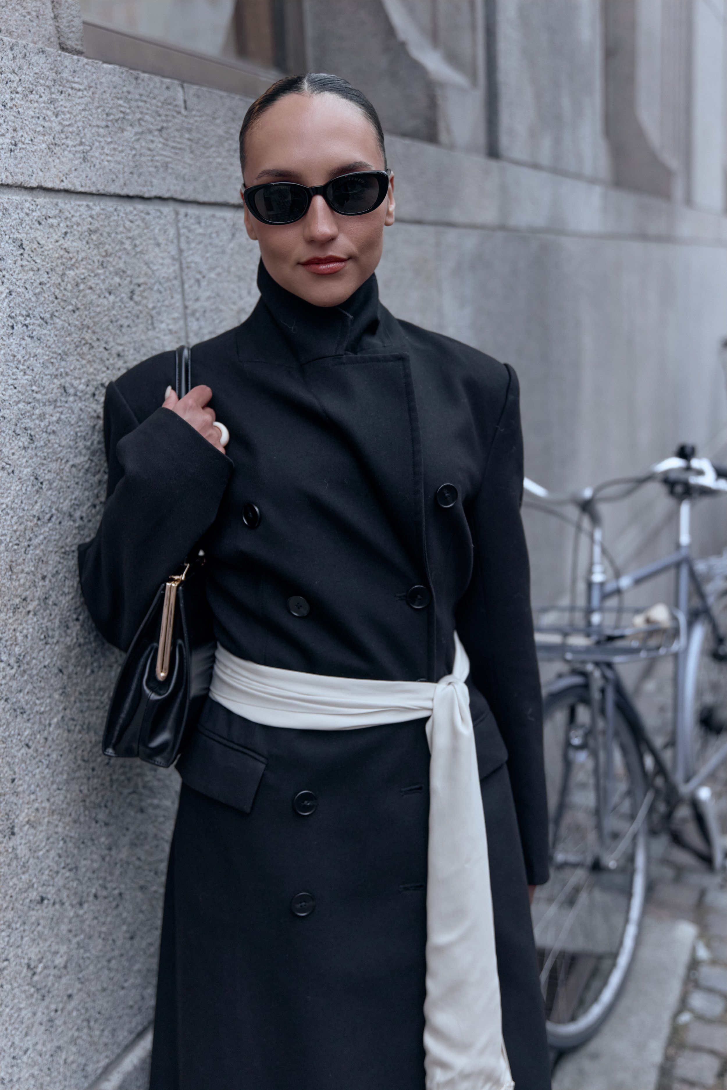 Woman in sunglasses and a black double-breasted coat tied with a white sash, bicycles behind her