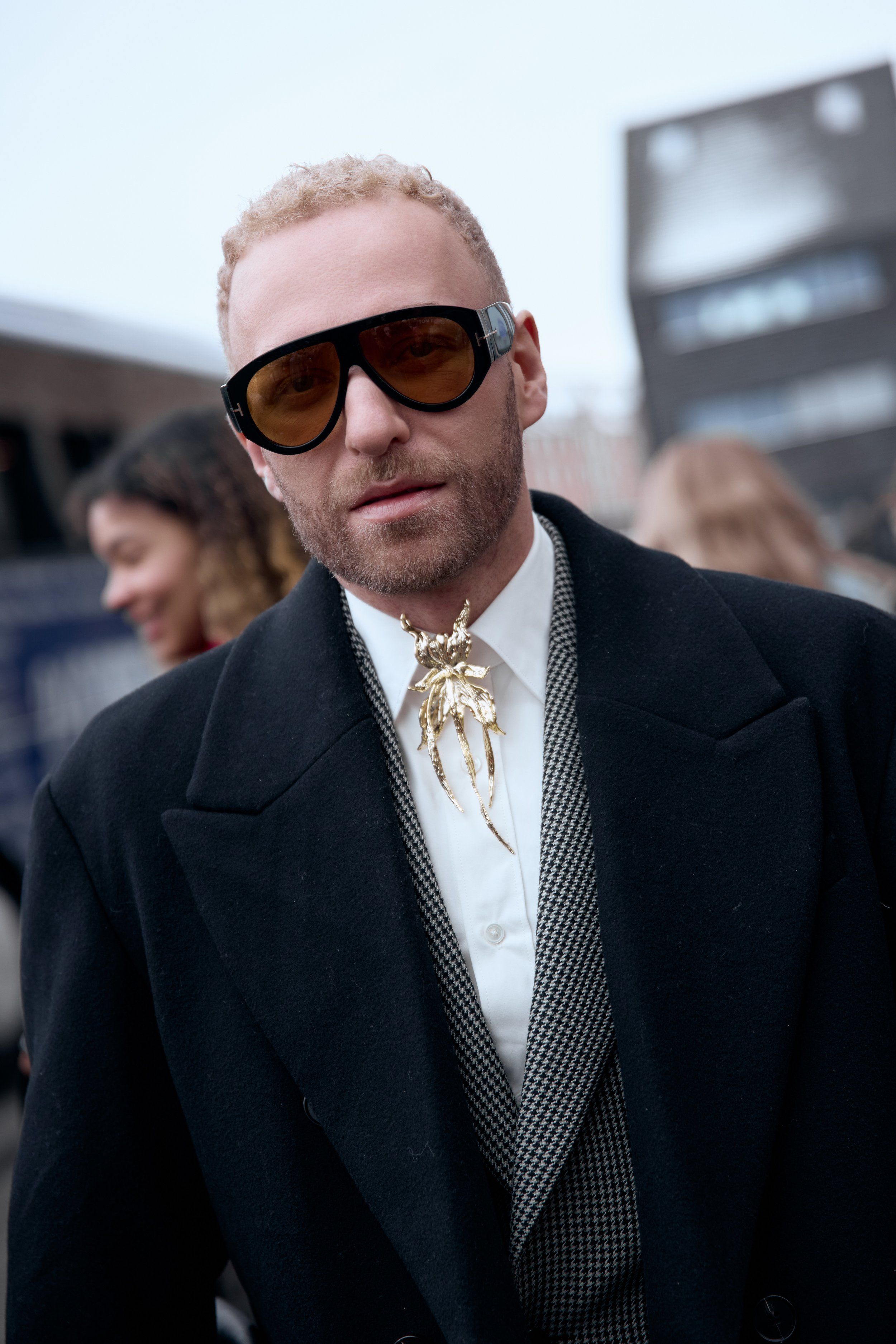 Man in tinted sunglasses and a black overcoat with a gold brooch on a white shirt and houndstooth waistcoat