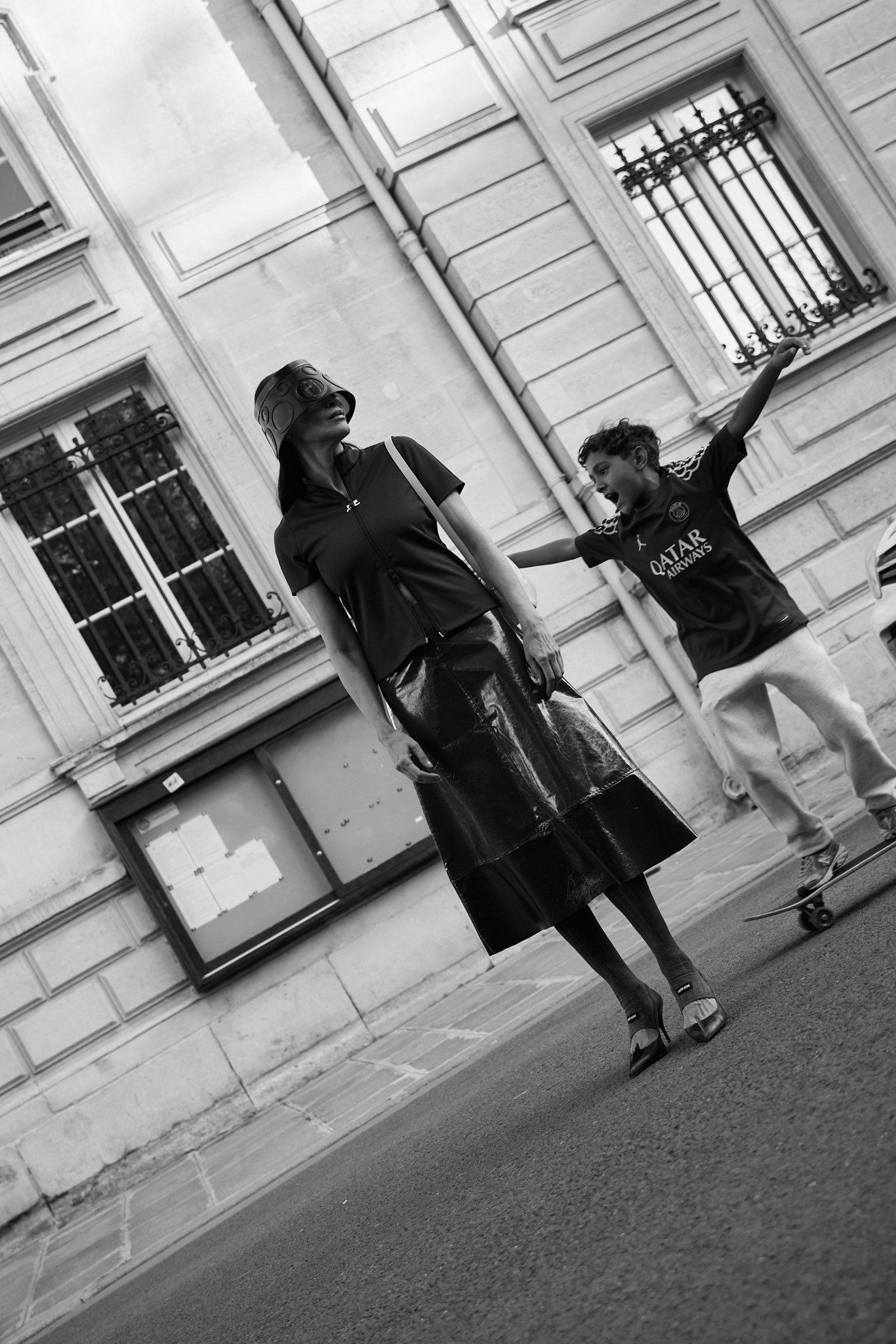Black-and-white shot of a woman in a leather skirt and a boy skating with arms raised on a city street