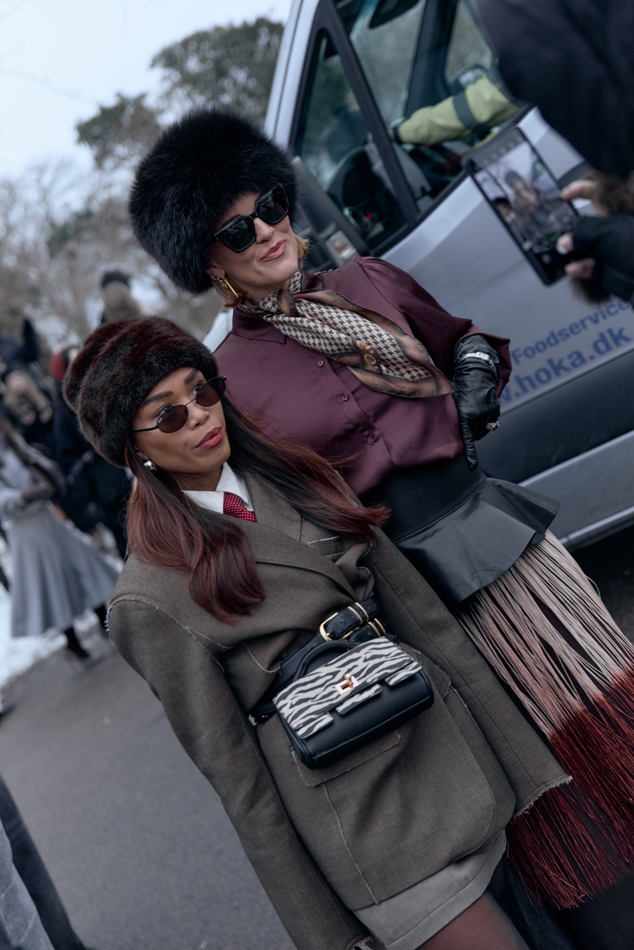 Two guests: one in fur hat and burgundy blouse, one in a brown blazer with a zebra-print shoulder bag