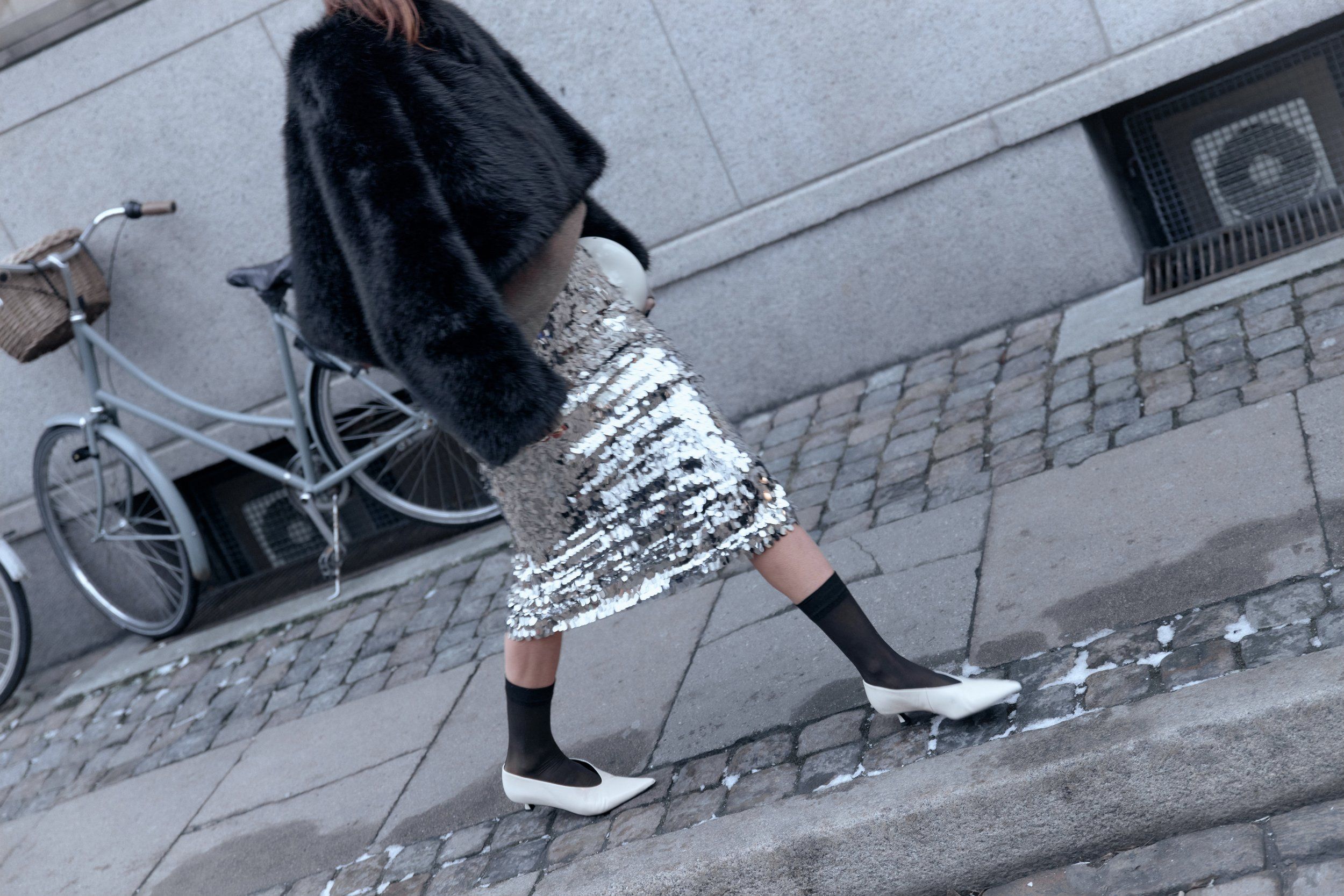 Woman in a black fur jacket and sequin skirt walking past a bicycle, white pointed shoes and black socks