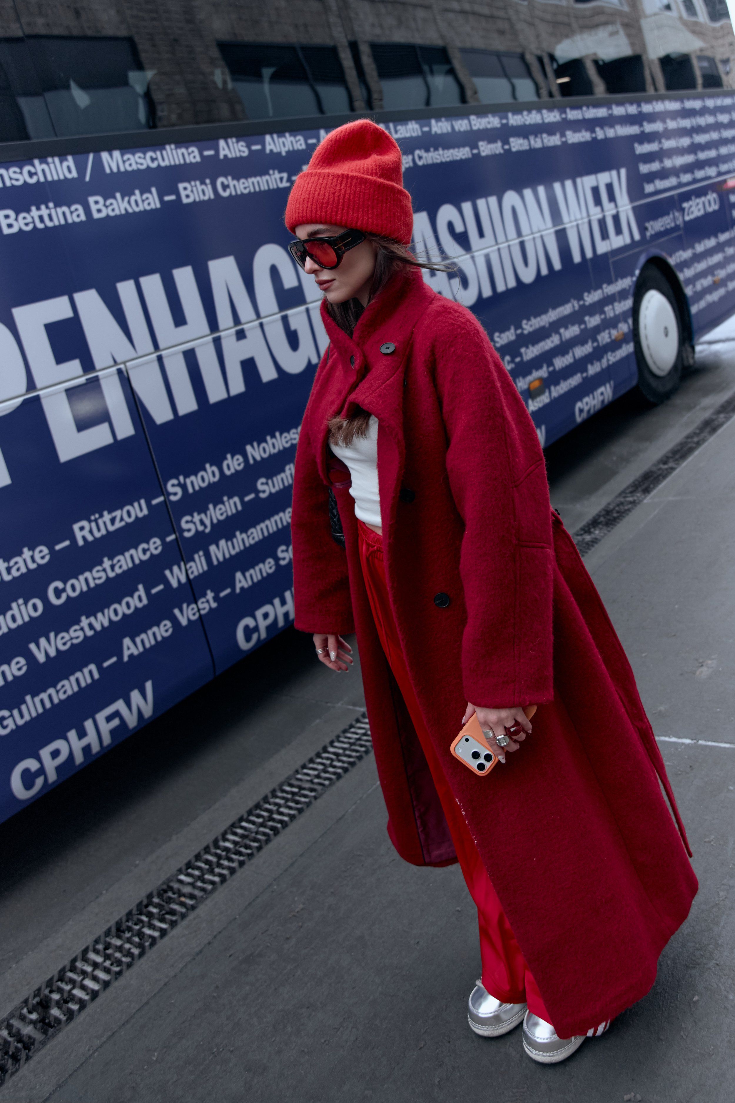 Woman in a red knit hat, long red coat, red trousers and silver shoes beside a CPHFW bus