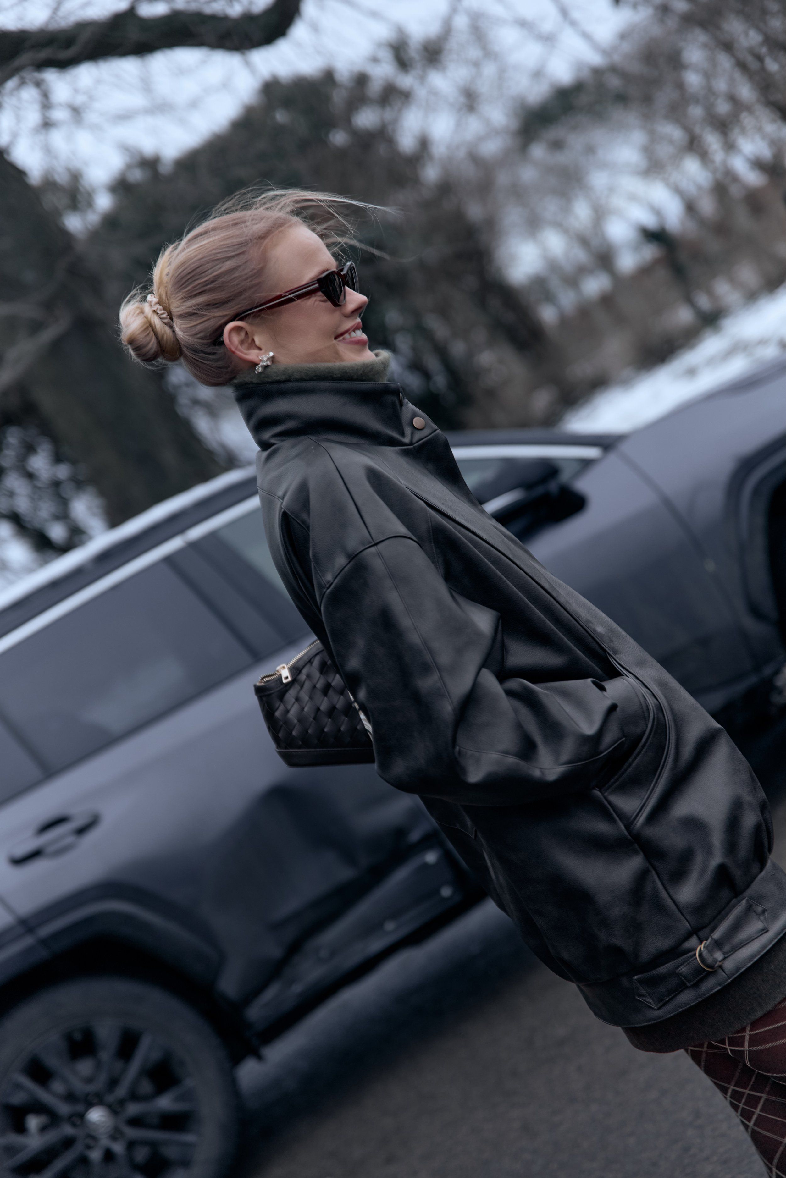 Woman in sunglasses with a low bun and an oversized black leather jacket, small woven bag, beside a car