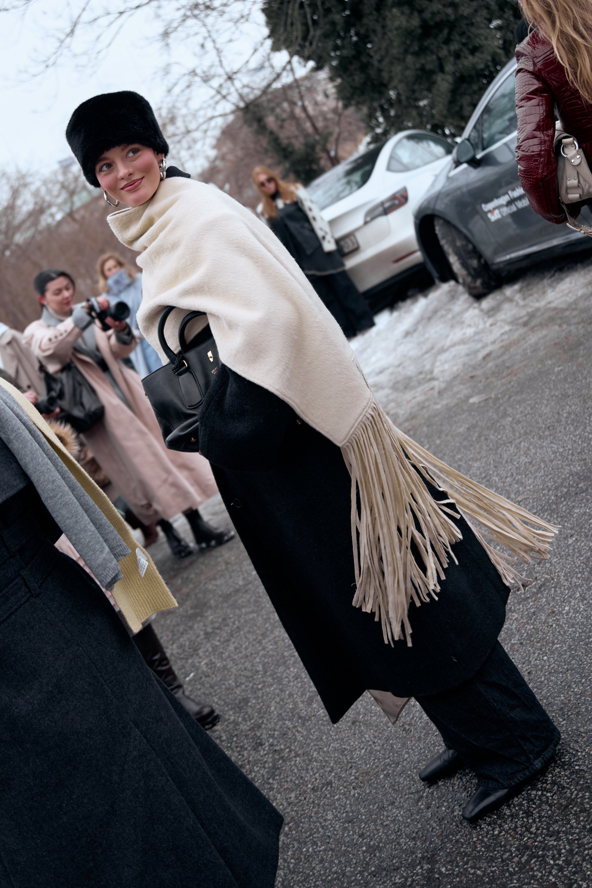 Woman in a black fur hat, cream shawl with long fringe and black trousers glancing back on a snowy street