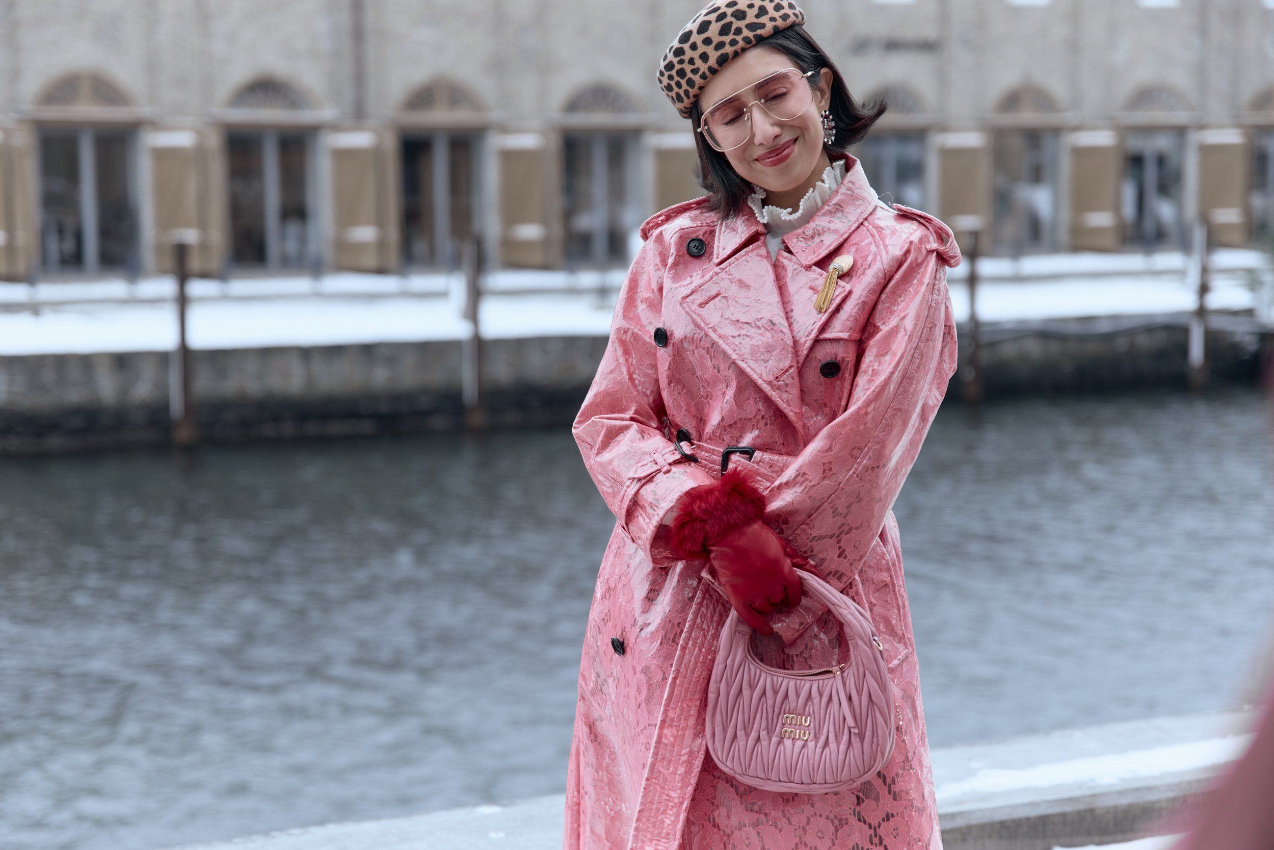 Woman in a pink glossy trench coat, red gloves and a leopard-print hat beside a canal