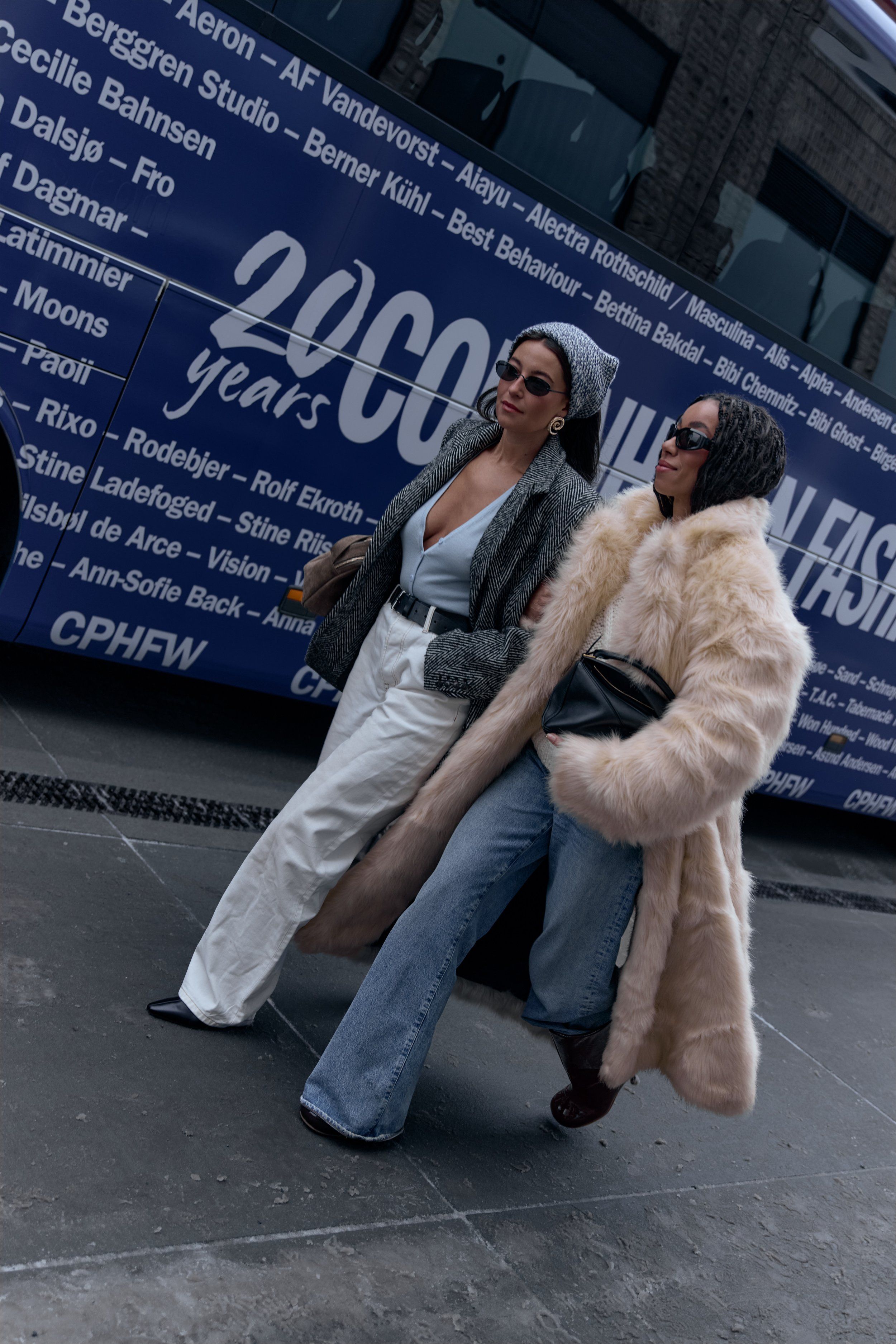 Two guests walking past the blue CPHFW bus, one in a cream fur coat, one in white trousers and grey knit