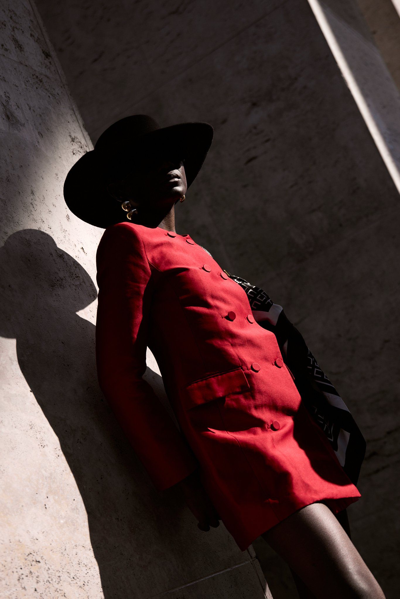 Woman in a red tailored jacket and black wide-brim hat posed against a stone wall in hard light