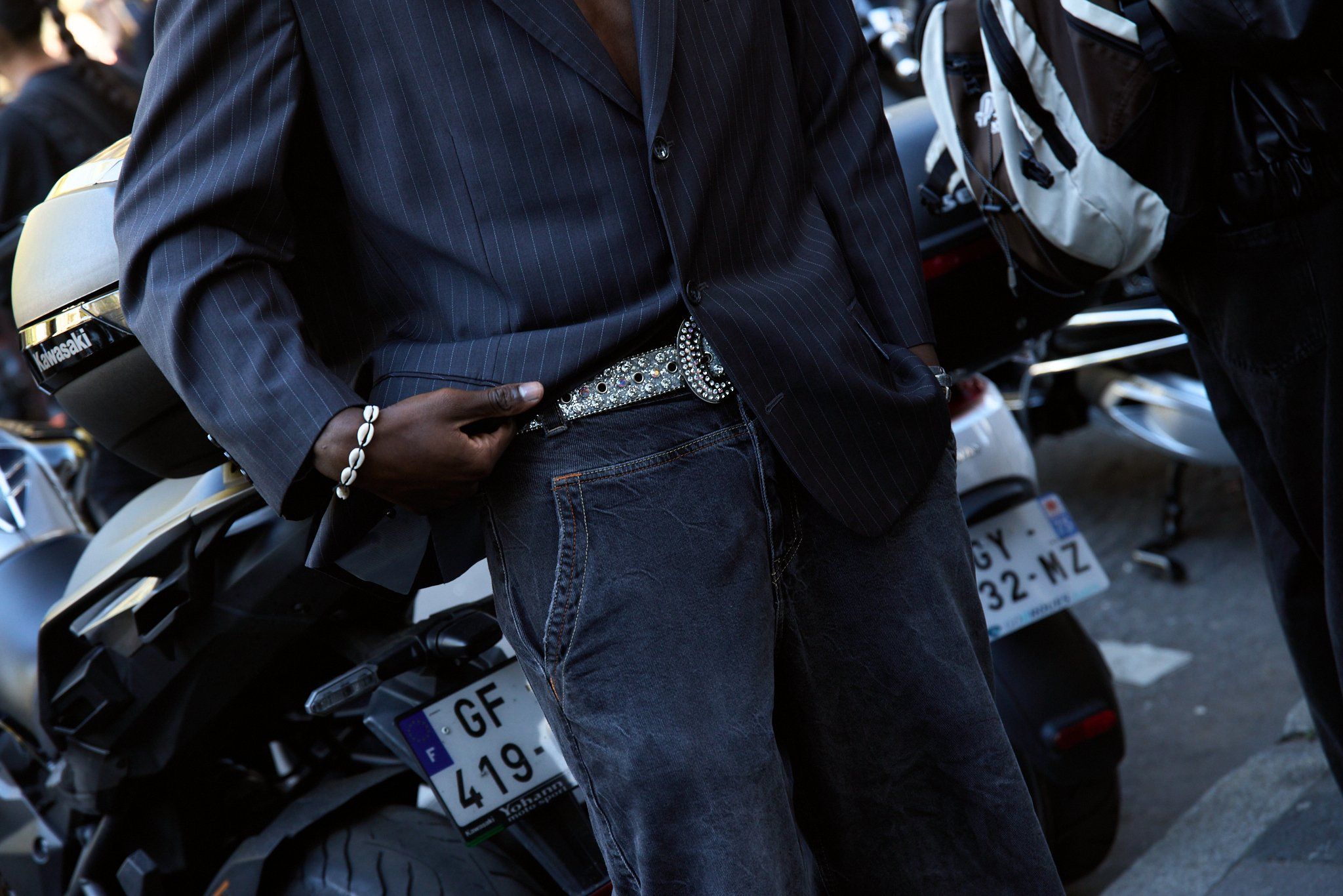 Man in a pinstriped dark blazer, jeans and a studded belt, motorbikes with plates behind him