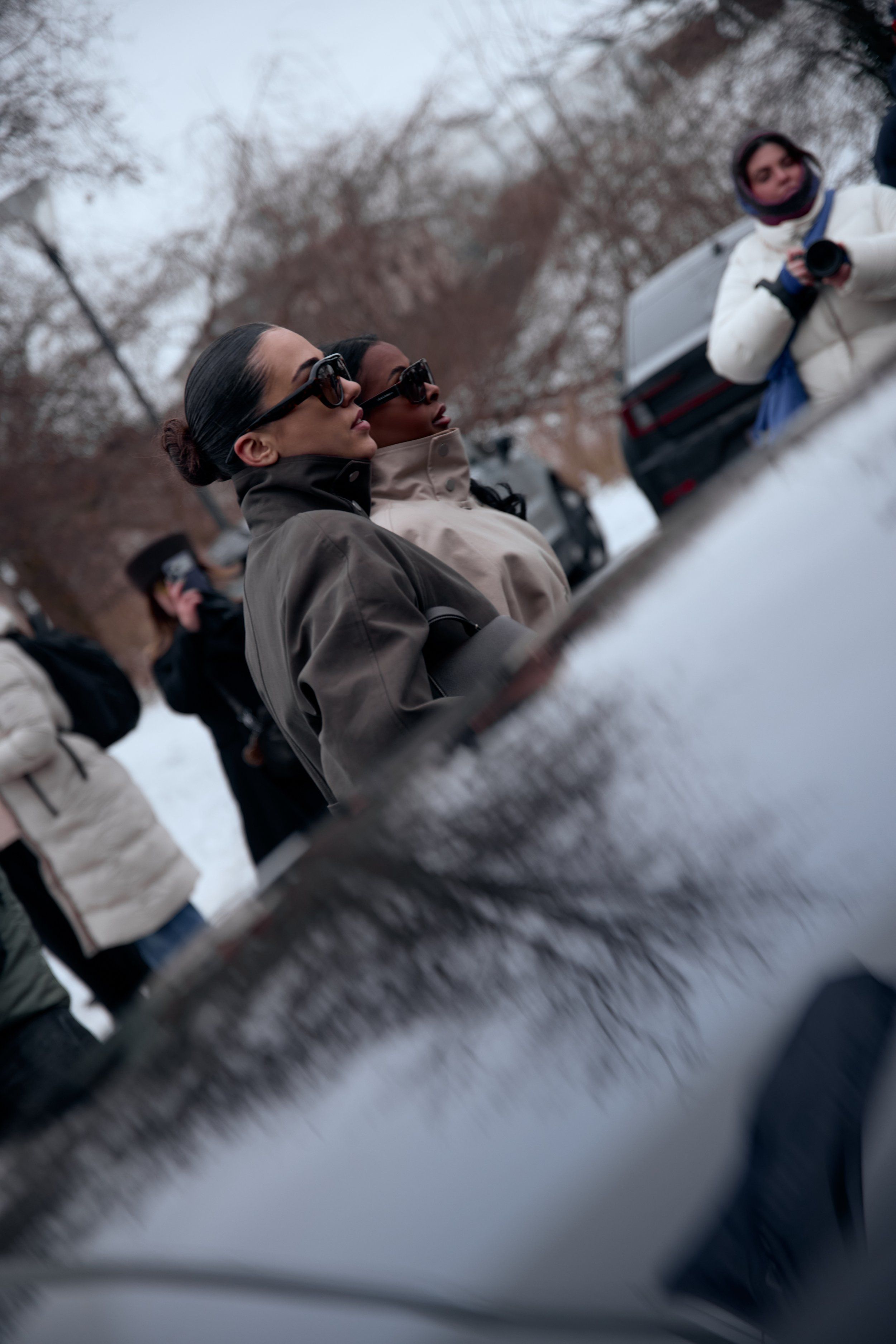 Guests in sunglasses beside a car roof reflecting bare trees, photographers behind them