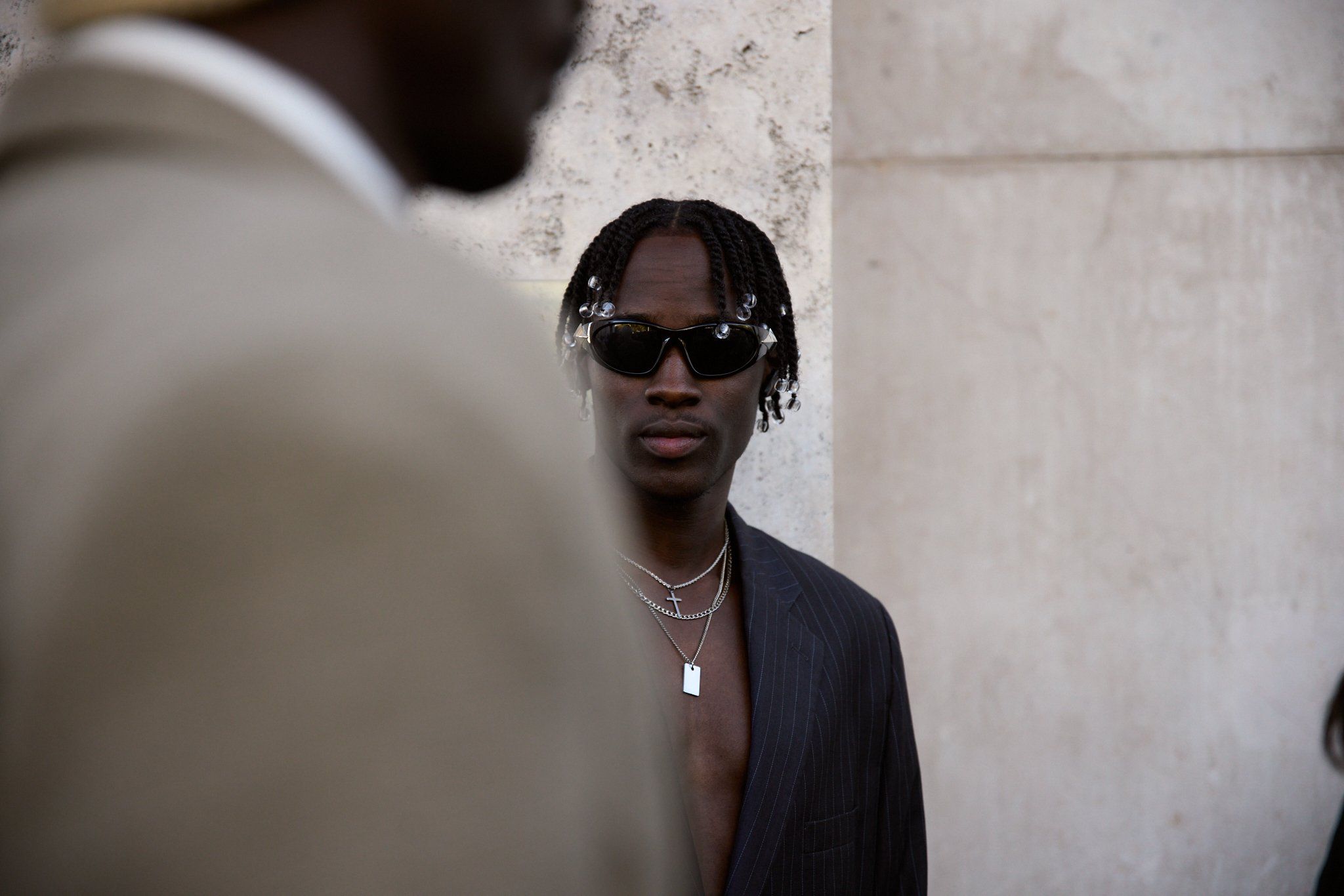 Man in dark sunglasses with braided hair and a grey blazer over bare chest, necklace chains, by a stone pillar