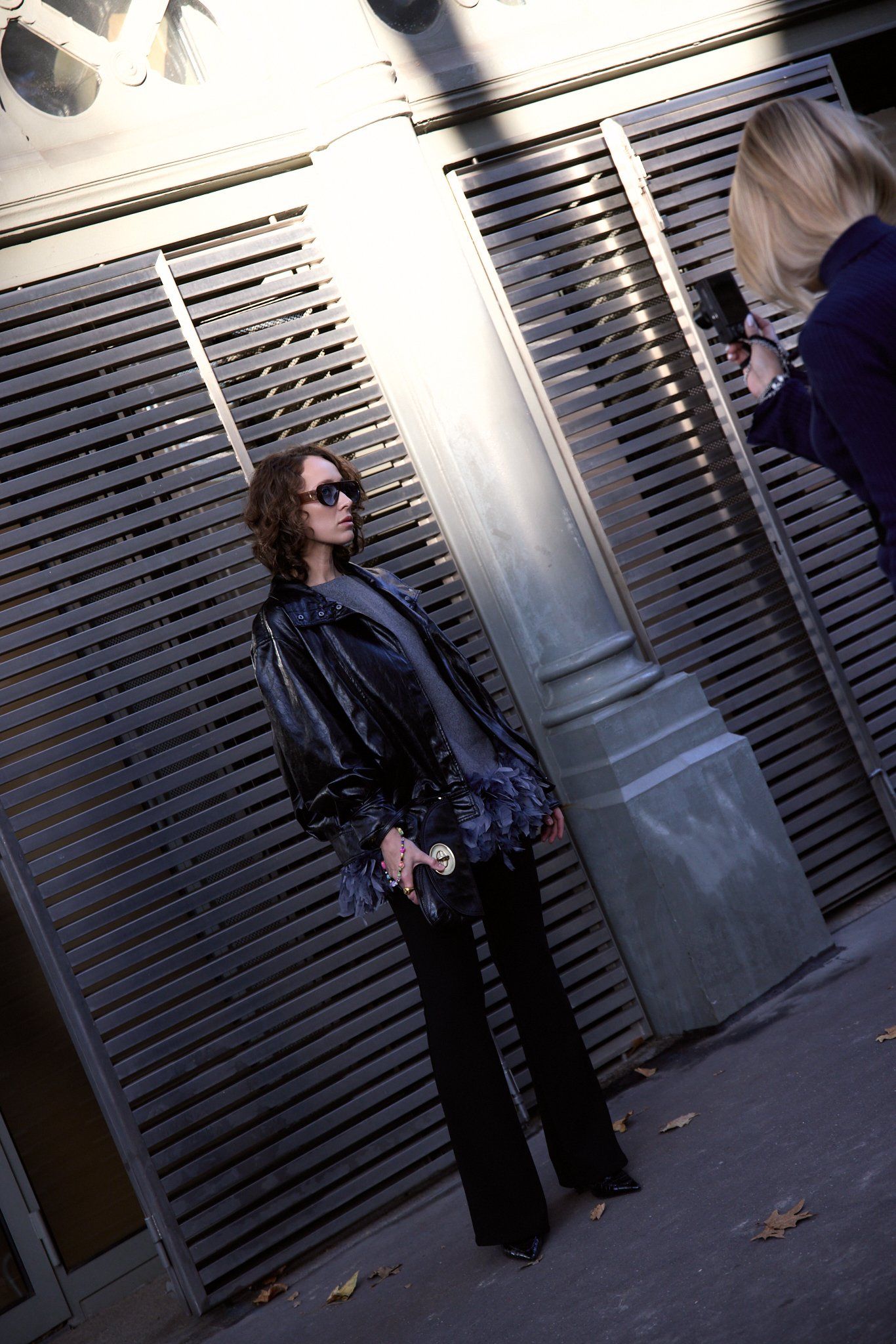 Woman in sunglasses and a glossy black jacket with feathered cuffs against metal shutters