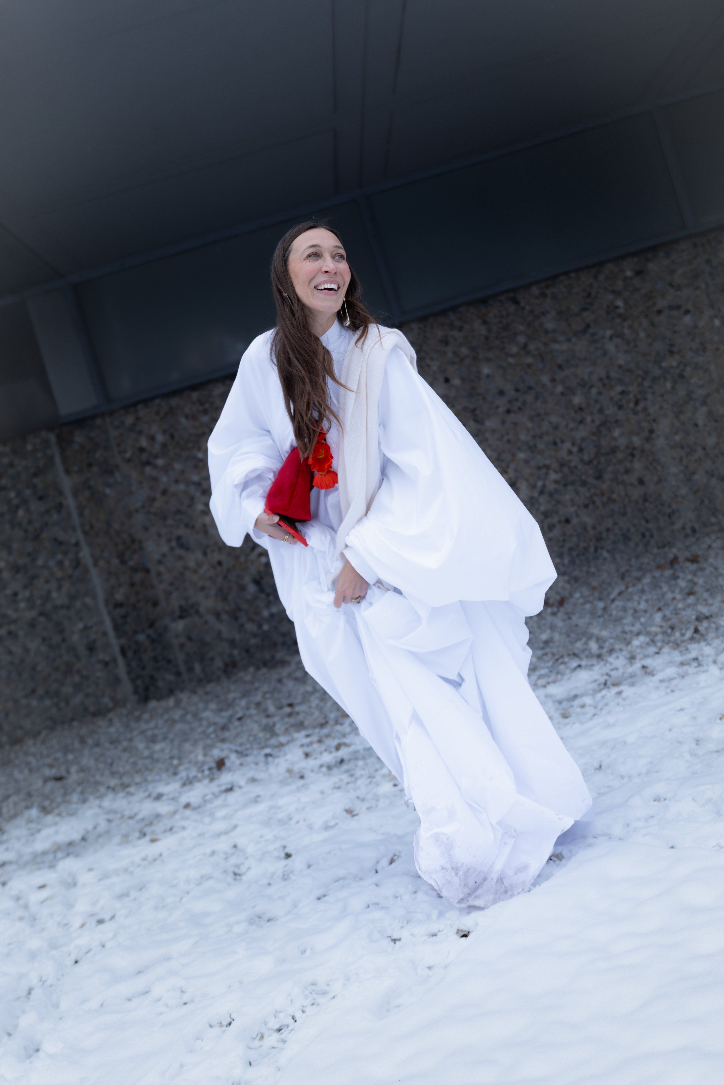 Woman in a white voluminous cape dress holding a red bag, laughing in front of a snowy ground