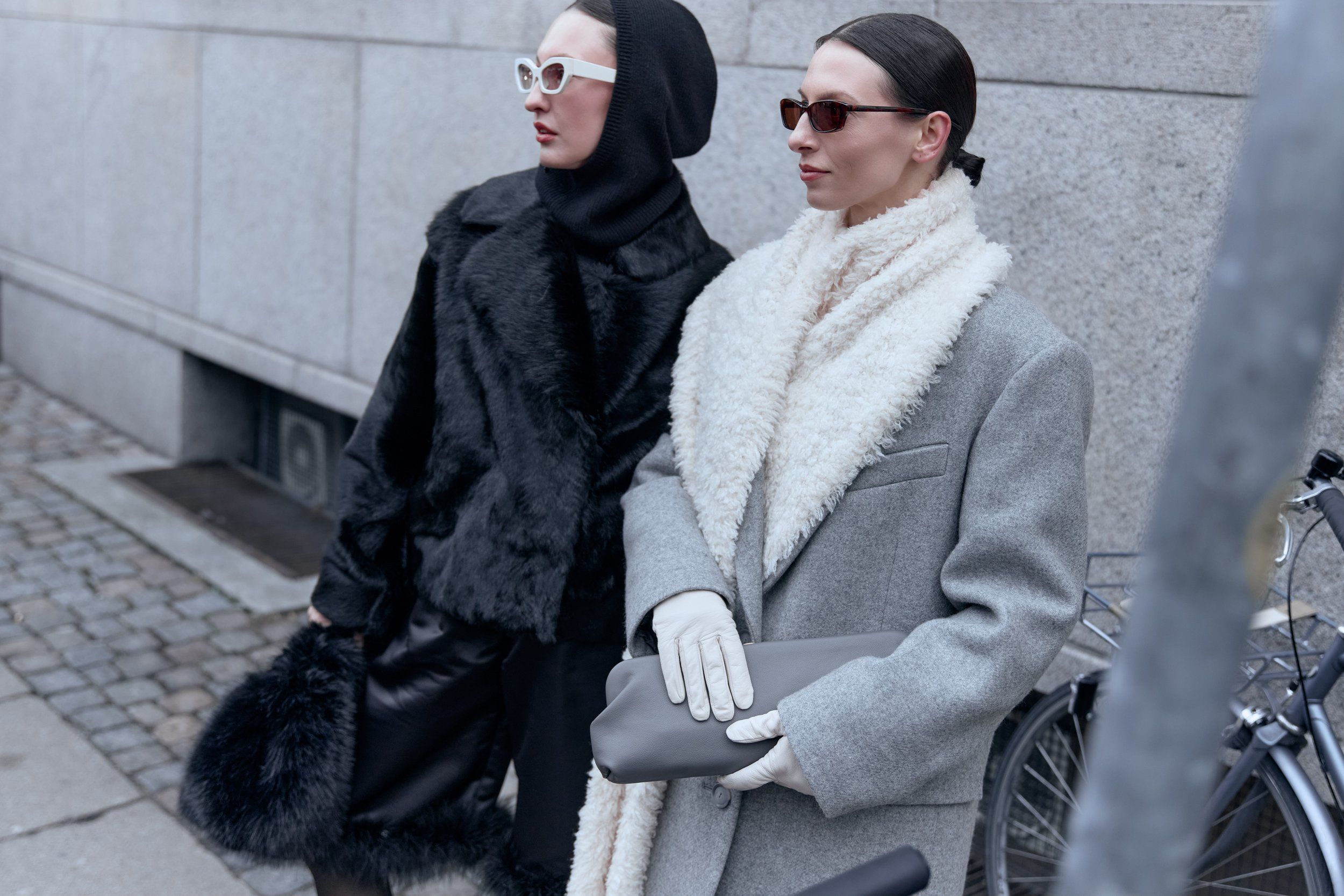 Two guests by a wall and bicycle, one in a black fur hood and one in a grey coat with white shearling collar