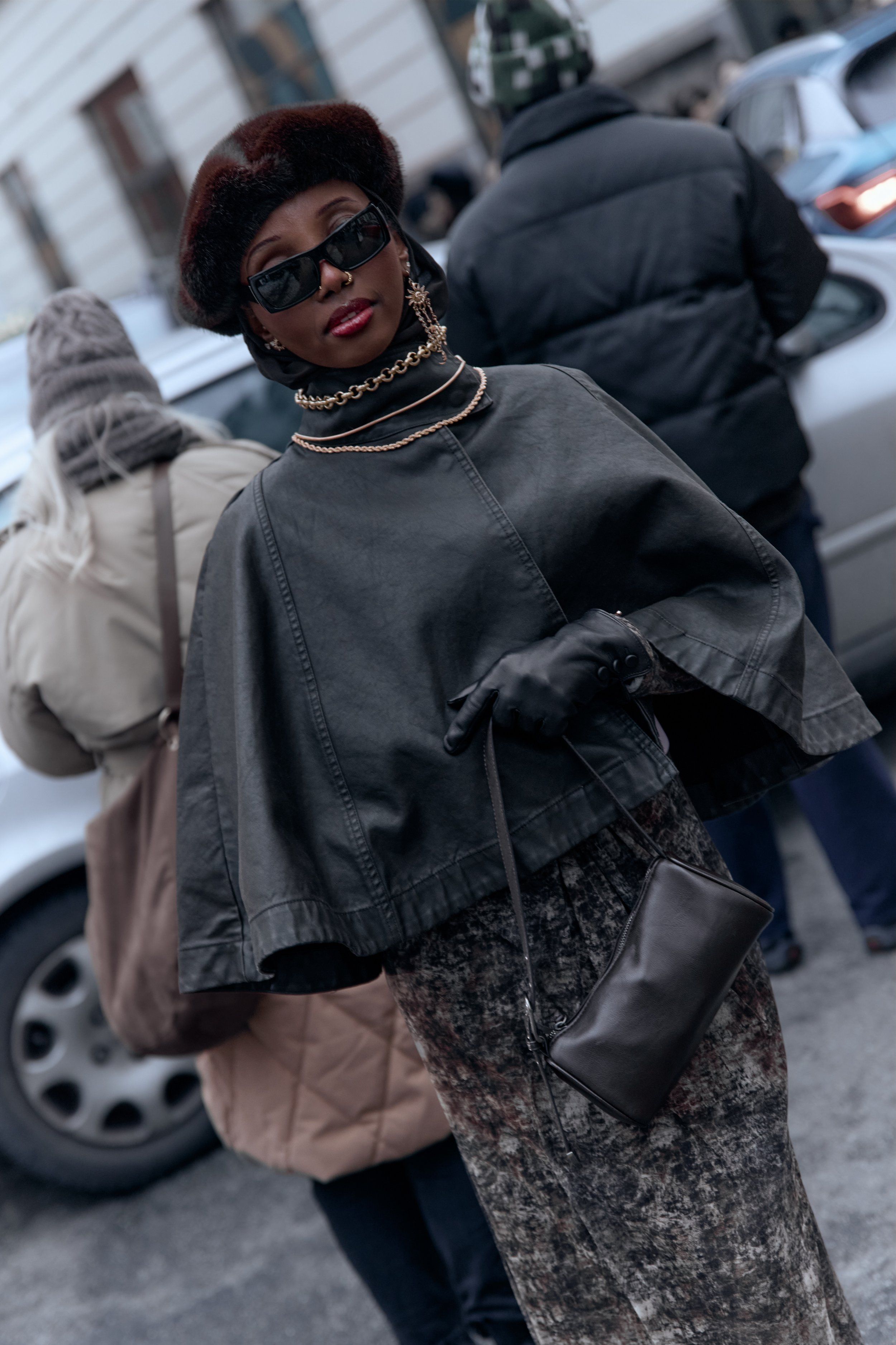 Woman in sunglasses and brown fur hat, a grey cape jacket, gloves and a mottled skirt with a small bag