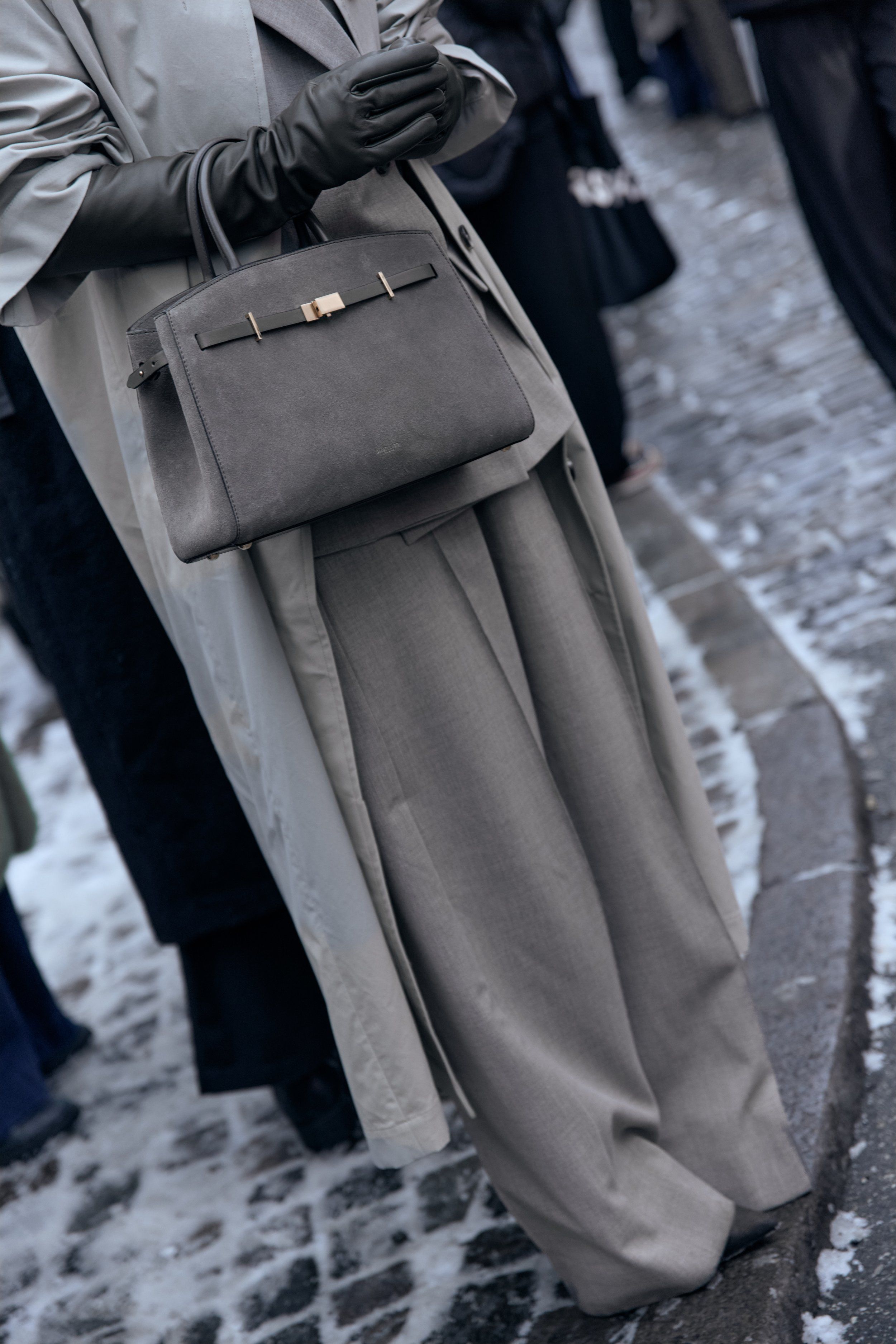 Grey trench coat, black gloves, wide grey trousers and a grey structured top-handle bag on a snowy cobbled street