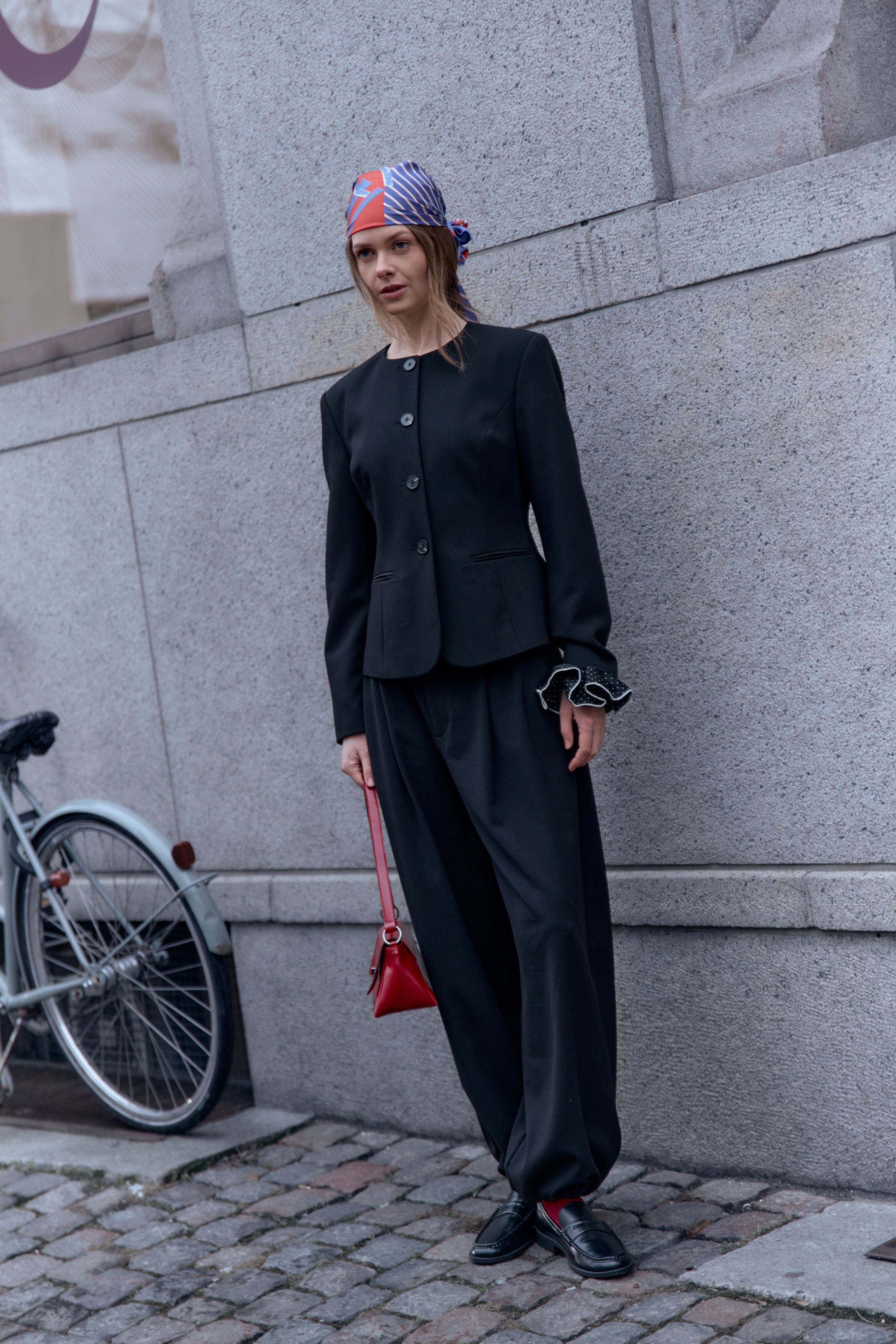 Woman in a black jacket and trousers with a patterned head scarf and small red bag, bicycle behind